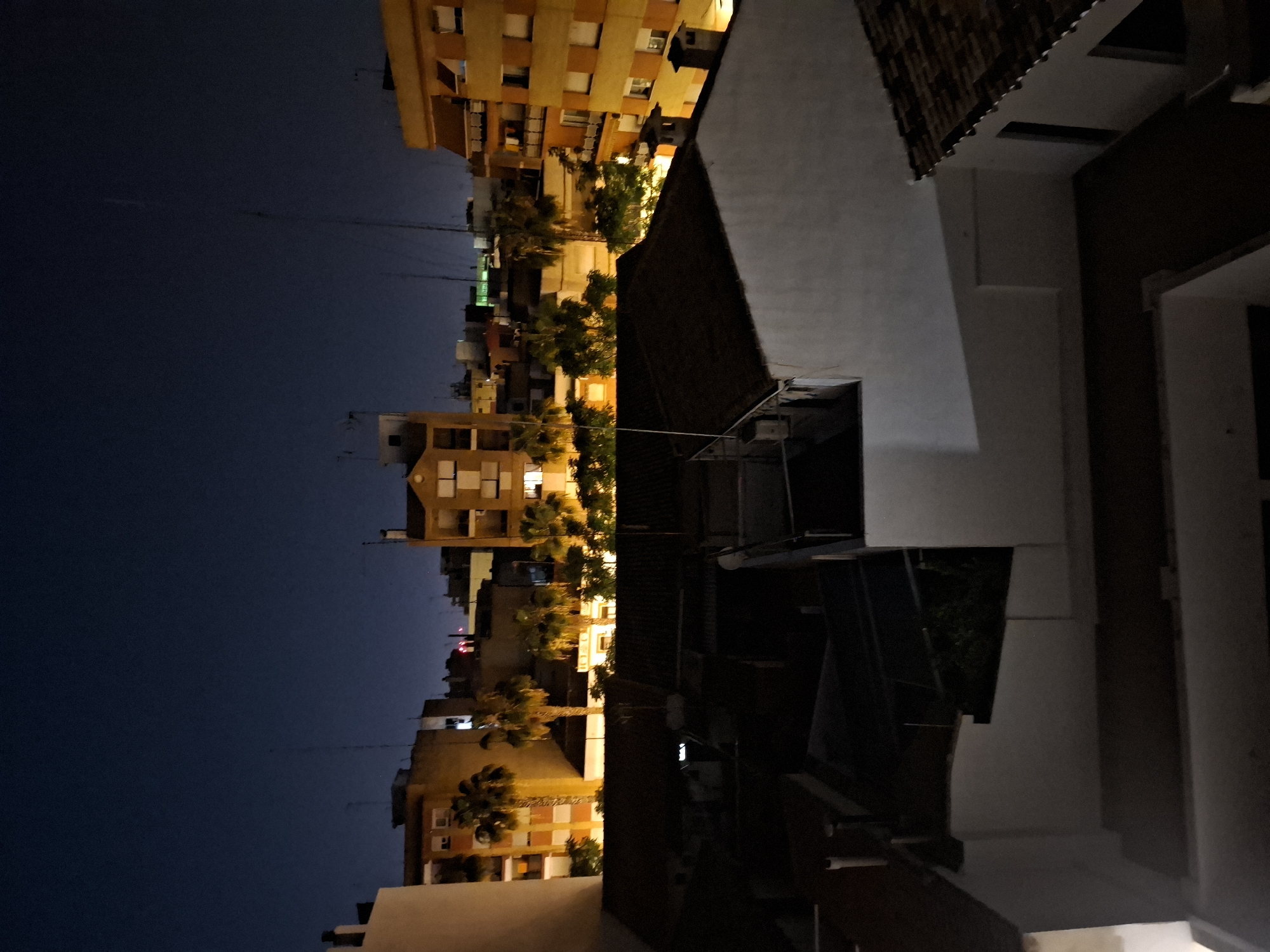 The image shows a night view of buildings in Xirivella, Spain. In the distance, a row of apartment buildings with lit windows stands against a dark sky. Palm trees line the street, adding to the urban landscape. In the foreground, portions of a closer building appear with white walls and dark roof tiles. The overall scene depicts an urban environment at night.