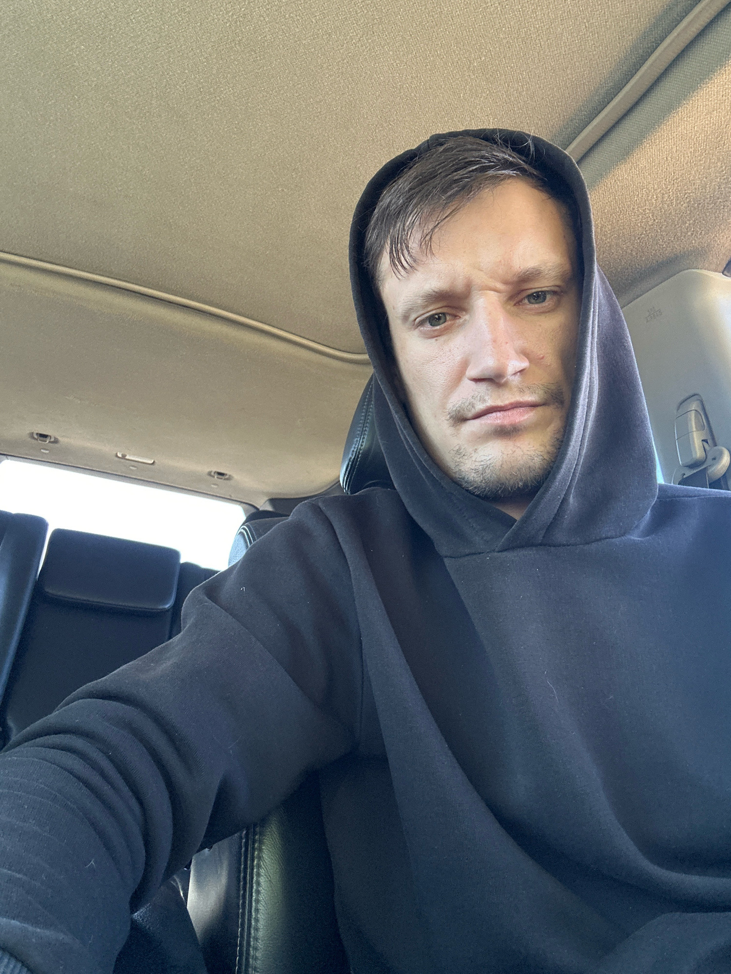 A man is sitting in the driver's seat of a car, wearing a black hooded sweatshirt. He is looking directly at the camera with a furrowed brow and a slight scowl. The interior of the car is visible, including the headliner, dashboard, and seats. The man's right arm is resting on his lap, and his left arm is bent at the elbow, with his hand resting on the steering wheel. The background is out of focus, but it appears to be a daytime scene. The man's clothing and the car's interior suggest a casual setting. There is no indication of the specific location within the city of Makiv, Ukraine, beyond the context of being inside a vehicle.