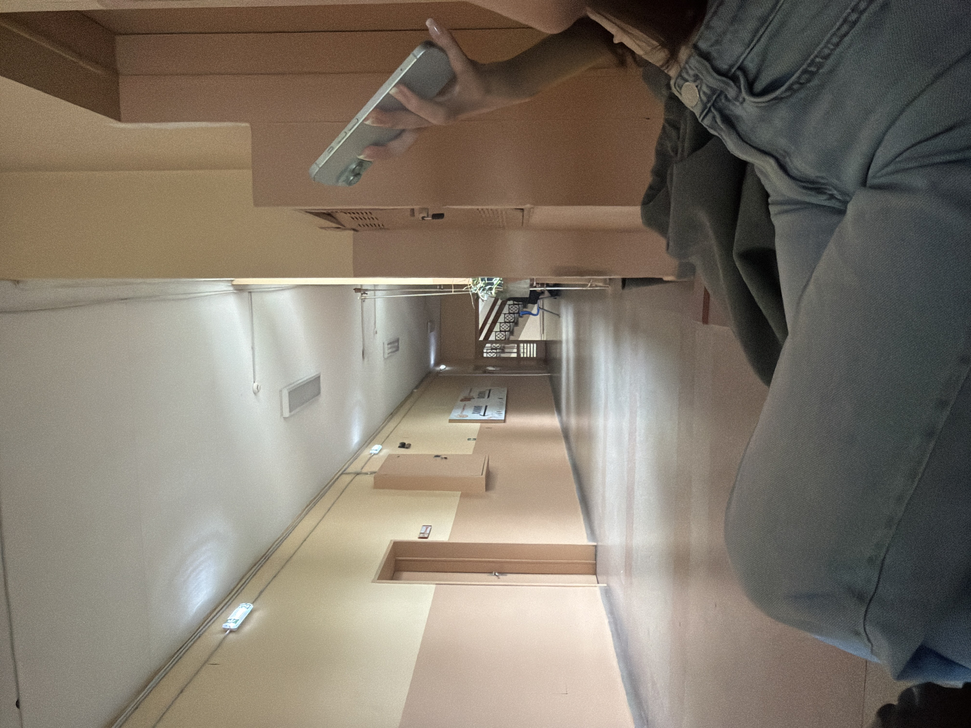 This image, taken in Sofia, Bulgaria, depicts a long, interior hallway, likely within a building, with a person partially visible on the right side. The hallway walls are painted in two horizontal sections: a lighter beige/cream on top and a peach-beige on the bottom. The floor also features a two-tone pattern of light beige and peach-beige sections.
On the right, a person is seen from the waist down, wearing light blue jeans and possibly a grey top. Their right arm is visible, holding a silver or light grey smartphone with a multi-lens camera module. The person appears to be standing or bending slightly, possibly taking a picture or observing their surroundings.
The hallway extends into the distance, where a dark metal staircase with railings is visible. Along the walls, there are several rectangular light fixtures and air vents. Near the bottom of the left wall, white wires run horizontally. Several rectangular objects, resembling boxes or enclosures, are placed on the floor, some open and some closed. One larger white rectangular board with faint, illegible text is also on the floor further down the hallway. Brown doors are spaced along the walls. The scene is illuminated by artificial lighting from the fixtures. The overall impression is that of a functional, possibly institutional, corridor.