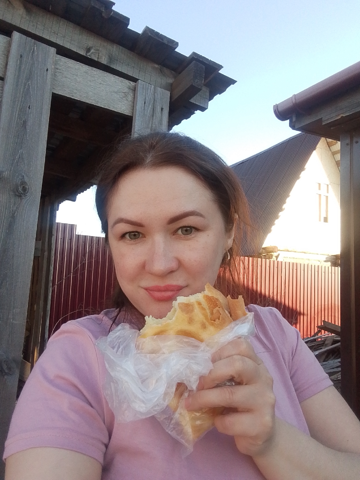 A woman is holding a piece of bread or pastry in a clear plastic bag. She is wearing a pink short-sleeved shirt. The background shows parts of buildings, including a fence, a wooden structure, and a house with a steeply pitched roof. The location appears to be in Прикамский, Russia, based on the wooden house structures.