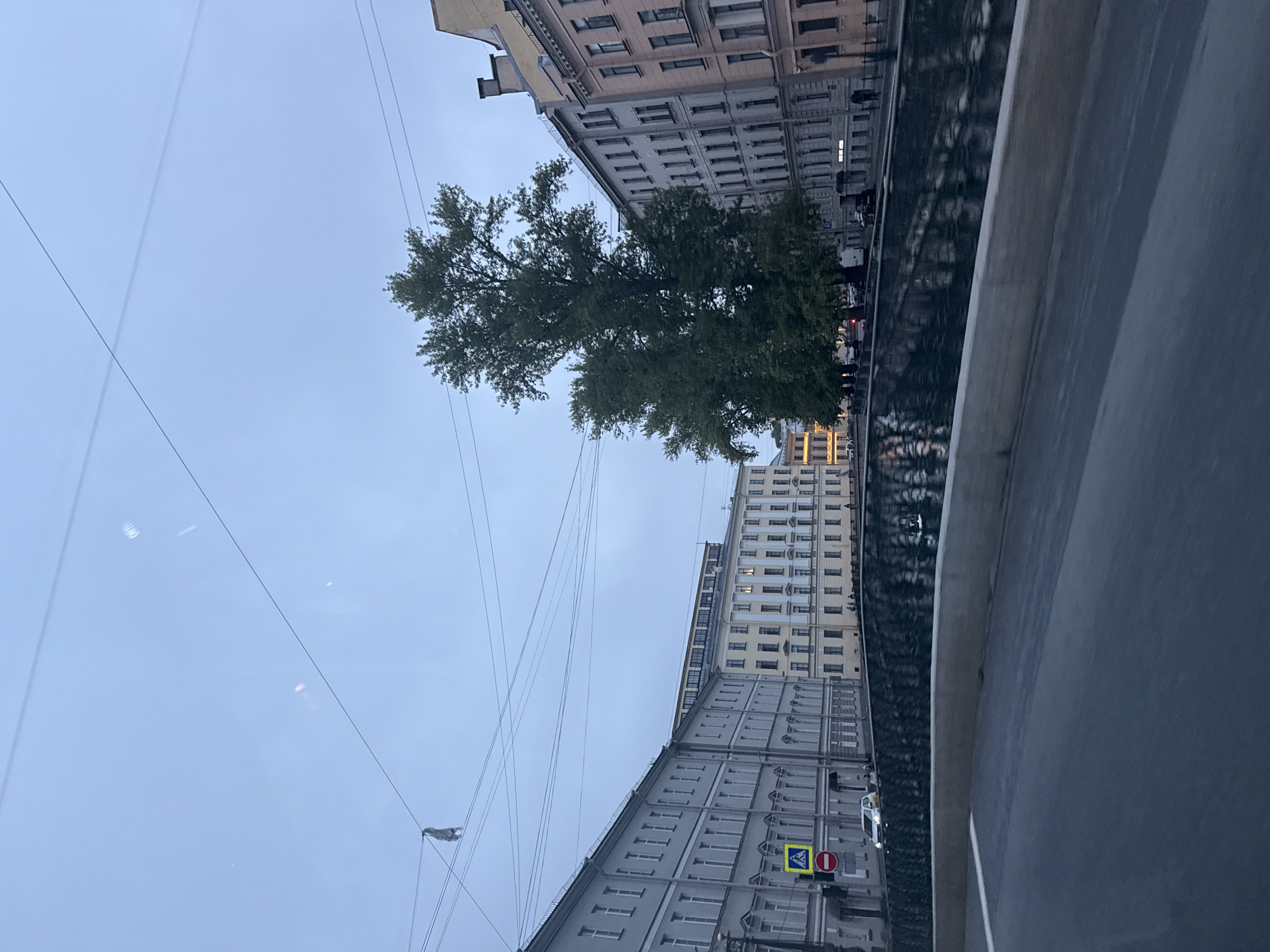 The image depicts a view of a canal and buildings in Saint Petersburg, Russia, from a slightly elevated perspective, possibly from a moving vehicle. The sky is overcast and a pale blue. Several wires crisscross the sky, and a grey, possibly fabric object is hanging from one of them on the left side of the frame.
The canal, lined with trees on the far bank, reflects the overcast sky and the buildings. The buildings are typical of Saint Petersburg architecture, with multiple stories, numerous windows, and ornate facades in shades of grey, beige, and pink. Some buildings have illuminated windows.
On the near bank of the canal, a dark, reflective surface suggests a bridge or embankment, with a concrete barrier in the foreground. Road markings are visible on this surface. A traffic sign with a yellow background and a blue pedestrian symbol, along with a red circular sign with a diagonal bar, are present on the sidewalk.
Several cars are visible on the street along the canal, and a few pedestrians can be seen walking. A large, mature tree with dense green foliage stands prominently on the far side of the canal, between the buildings and the water. The overall atmosphere is calm and somewhat subdued, consistent with a cloudy day in an urban environment.