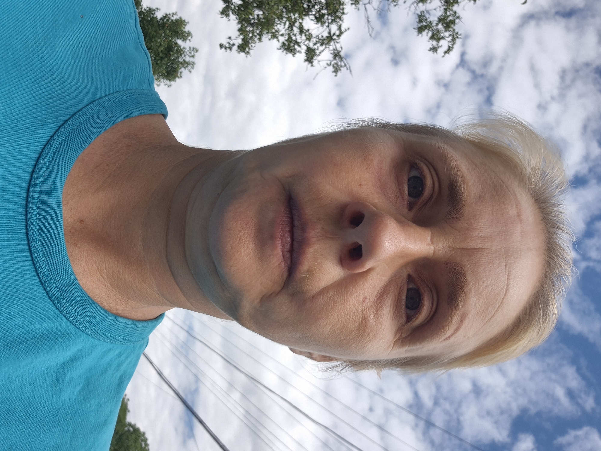 A woman with fair skin and blonde hair stands outdoors, facing the camera. She is wearing a bright blue t-shirt. Her eyes are open, and her mouth is slightly parted. The background consists of a cloudy blue sky, with trees and power lines visible. The image appears to be a selfie, taken from a low angle. The location is the city of Sozonivka, Ukraine.