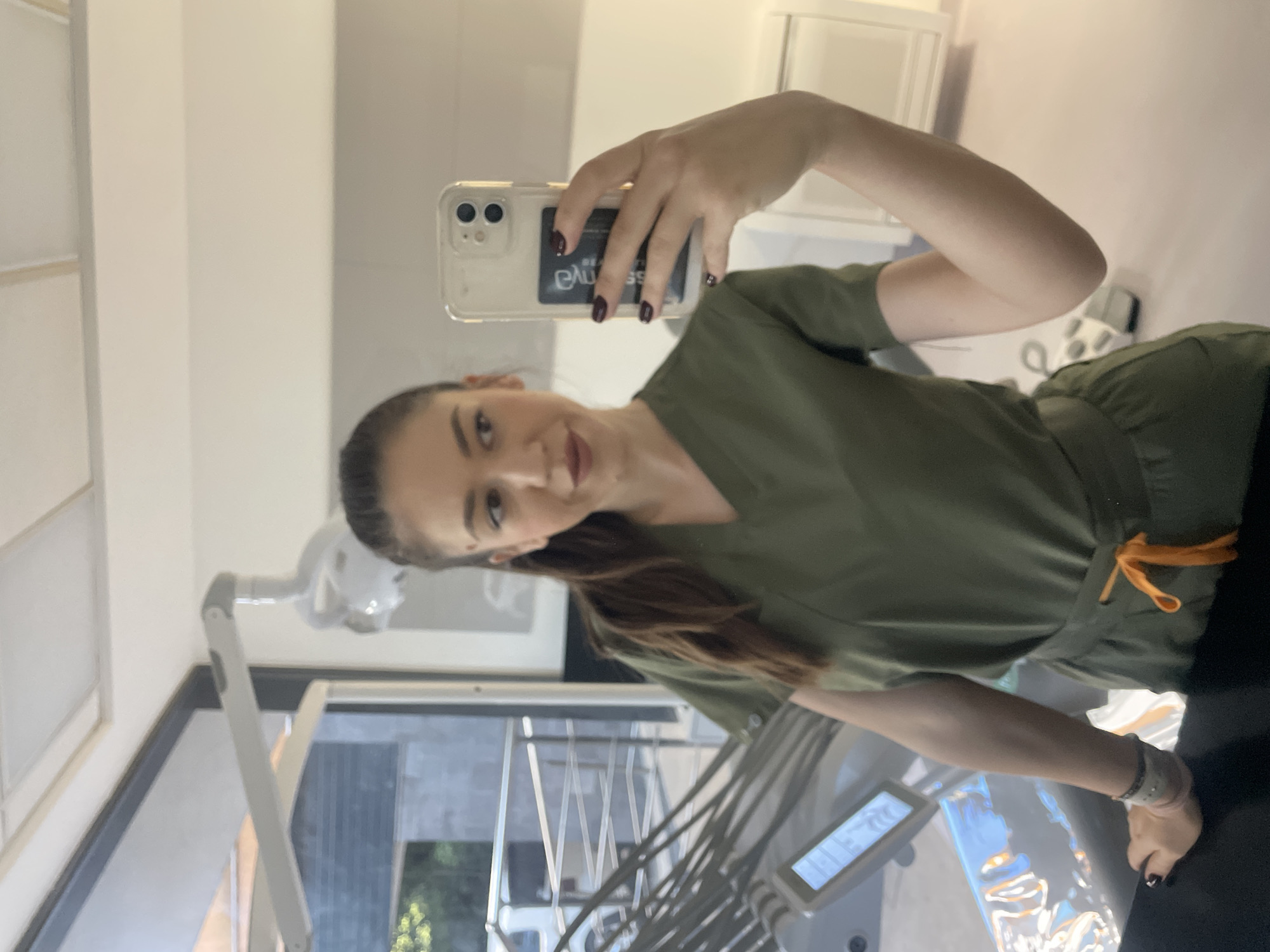 A young woman in a green scrub suit is taking a selfie in what appears to be a dental office. She is holding a smartphone in her right hand, with dark nail polish visible. Her hair is pulled back in a ponytail. Behind her is a dental chair, complete with various dental tools and equipment. The background includes a white wall and window. It is impossible to confirm the location as Sofia, Bulgaria.