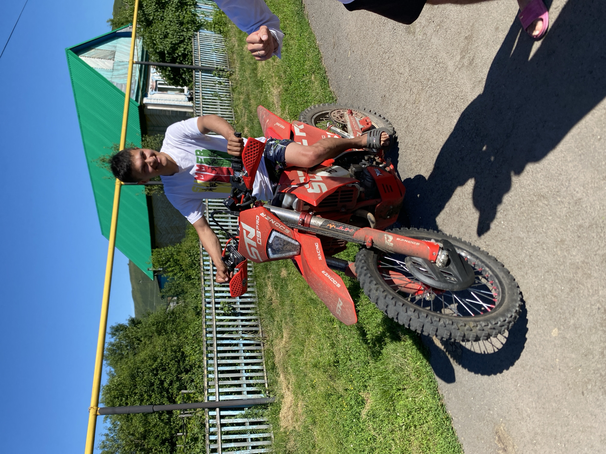 A young man is sitting on a red dirt bike in an outdoor setting. He is wearing a white t-shirt with a graphic design and dark shorts. His right arm is resting on the handlebars, and his left hand is gripping them. He is looking towards the camera with a slight smile.
The dirt bike is predominantly red with some black accents. The front wheel is visible, as is part of the rear wheel. The bike has knobby tires suitable for off-road use. There is visible text on the bike, including "RACING" and "SOONER".
The scene appears to be a sunny day, indicated by the bright sunlight and the clear blue sky. The background consists of green grass, a wooden fence, and a building with a green corrugated roof. There is also a yellow pole that appears to be part of a fence or utility line. The ground is a mix of asphalt and grass. The presence of a building and a fence suggests a rural or suburban environment, possibly a residential area. The overall mood seems relaxed and casual. The image was taken in Maloarslangulovo, Russia.