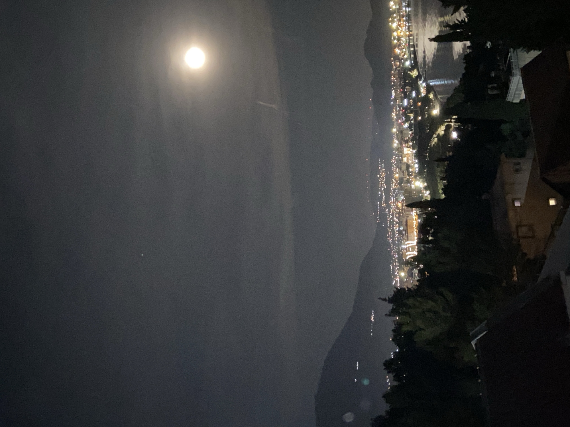 The image shows a nighttime view of Šušanj, Montenegro, with the moon visible in the upper left. A line of city lights stretches along the coast, separating the dark sea from the silhouette of mountains. In the lower right, a dark rooftop and foliage suggest an elevated vantage point. There are no people visible in the frame.