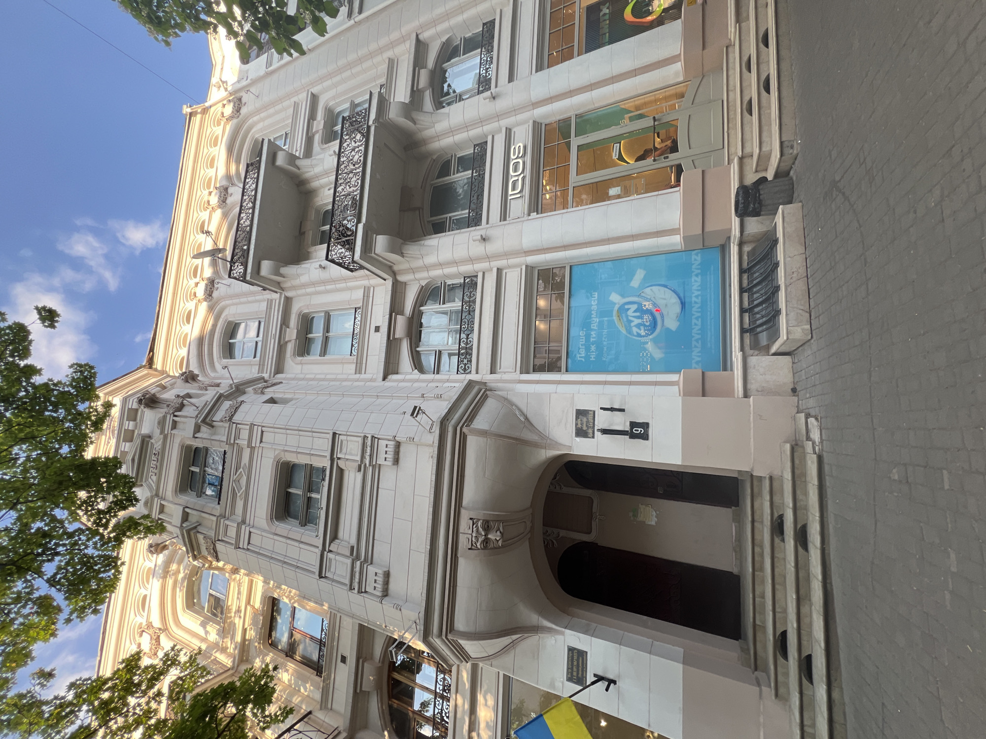 This is a vertical, eye-level, full-shot photograph of a building in Odesa, Ukraine. The building is light-colored and features intricate architectural details, including ornate balconies and decorative window surrounds. The sunlight casts a warm glow on the facade, suggesting it is daytime, likely late afternoon or early evening given the golden light. Trees with green foliage frame the left side of the building, and a clear blue sky with a few wispy clouds is visible at the top.
The building appears to be a commercial establishment, with several storefronts visible. One prominent sign reads "IQOS," and another large blue banner displays the "ZYN" logo, along with text in Ukrainian that translates to "Easier than you think." There's also a hint of a shop with brightly lit displays and a person inside, visible through a glass window. A Ukrainian flag is partially visible in the lower right corner. The street in front of the building is paved with cobblestones. The overall scene conveys a sense of urban life and commerce in a historic European city.