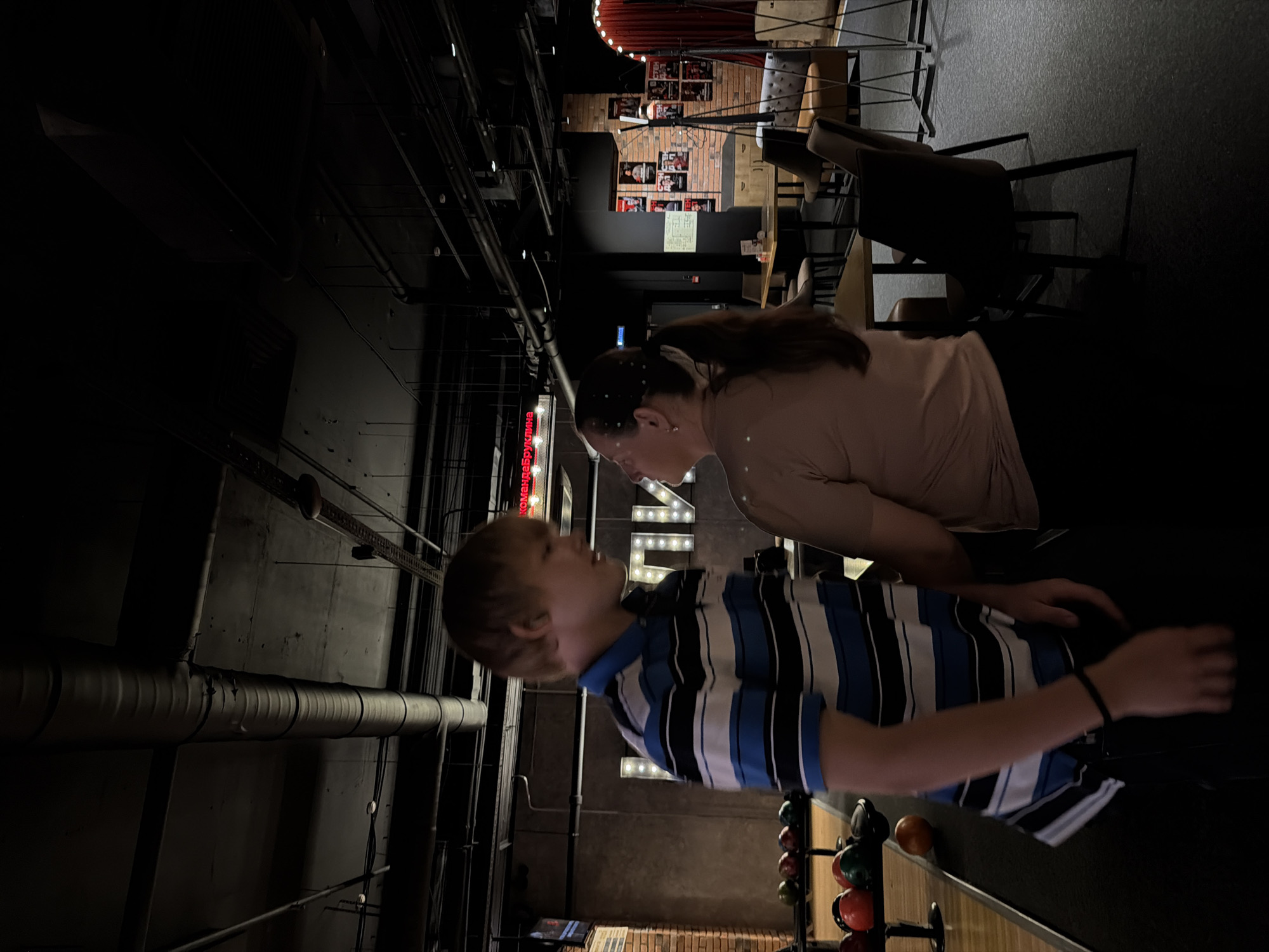 In a dimly lit bowling alley, a young man with blond hair, wearing a blue, white, and black striped polo shirt and dark pants, stands to the left, facing a woman. The woman, with dark hair pulled back in a ponytail and wearing a tan t-shirt and black pants, stands to the right, facing the young man. They appear to be interacting, with their faces close together. The background shows bowling lanes, balls, and seating areas. Above, industrial-style ductwork and lighting are visible. The scene is located in Izhevsk, Russia.