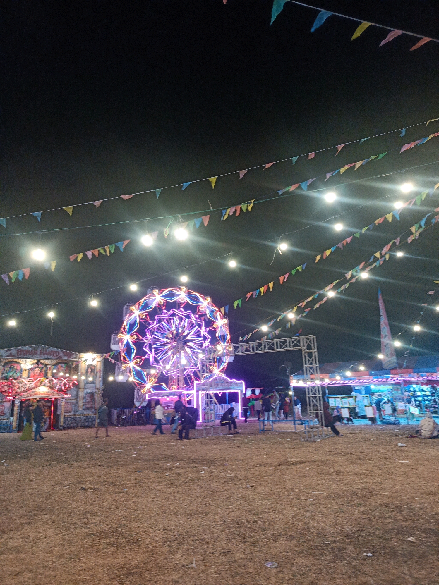The image depicts a night scene at what appears to be a local fair or carnival in Tanjungsari, Indonesia. A large, brightly lit Ferris wheel with intricate neon lights in purple and blue dominates the central area. Colorful triangular flags are strung across the sky, interspersed with bright lights, creating a festive atmosphere.
To the left of the Ferris wheel, a stall or attraction with neon lighting and signage is visible, featuring illuminated images. Several people are present, some walking around the grounds, while others are near the attractions. The ground is covered in dry grass or straw, typical of an outdoor event space. In the background, additional structures and lights suggest a variety of stalls and entertainment options. The overall impression is a lively community gathering at night.