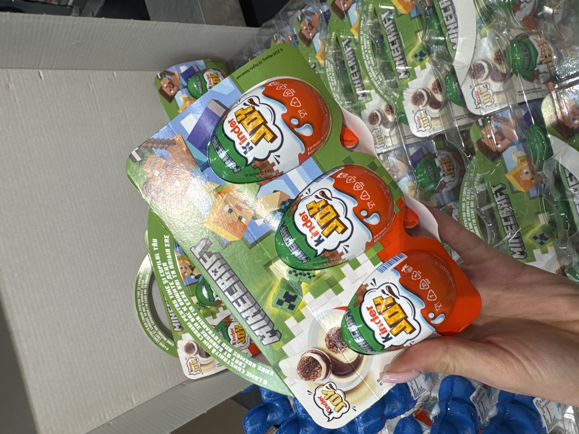 The image shows a hand holding a package of Kinder Joy Minecraft edition chocolate eggs. The packaging is predominantly green and features Minecraft imagery. A cardboard box is partially visible on the left. Multiple packages of the same Kinder Joy product are stacked behind the one being held. The image seems to be taken indoors under bright lighting conditions. There's no information available about the city of Varna, Bulgaria, or any specific location.