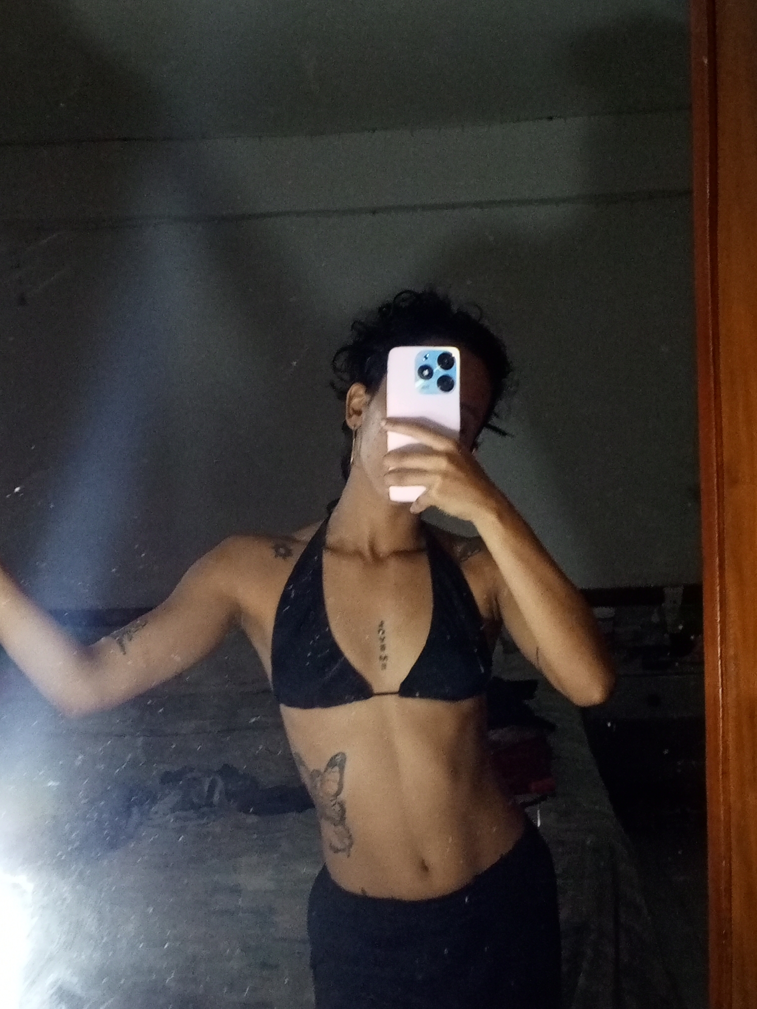 A person is taking a selfie in front of a mirror. The person is wearing a black bikini top and dark pants. The person is holding a pink phone in their right hand, covering their face. The person has tattoos on their left arm, chest, and left side. The mirror appears to be dirty. The location is likely indoors, with a simple wall in the background. The person is standing in front of what appears to be a bed. I cannot confirm the location of Porlamar, Venezuela.