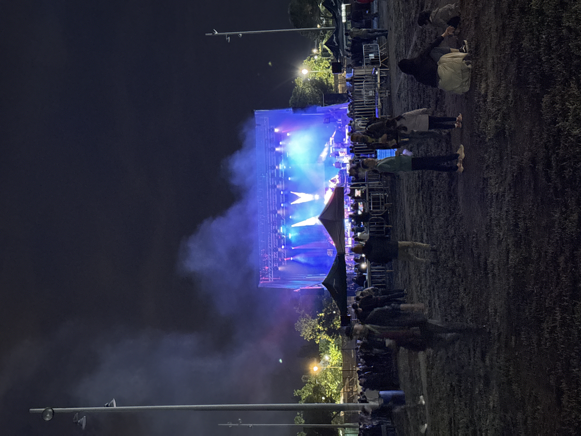 The image depicts an outdoor concert at night, with a stage illuminated by blue and white lights. A crowd of people is gathered in front of the stage, some standing and some sitting. A large cloud of smoke or fog is visible around the stage area. There are trees and streetlights visible in the background. The location is likely in Vilanova i la Geltrú, Spain, based on the provided information. There are tall light posts on the left side of the image. The crowd of people is mostly focused on the lit-up stage. Metal barricades are visible at the front of the stage, separating the audience from the performance area.