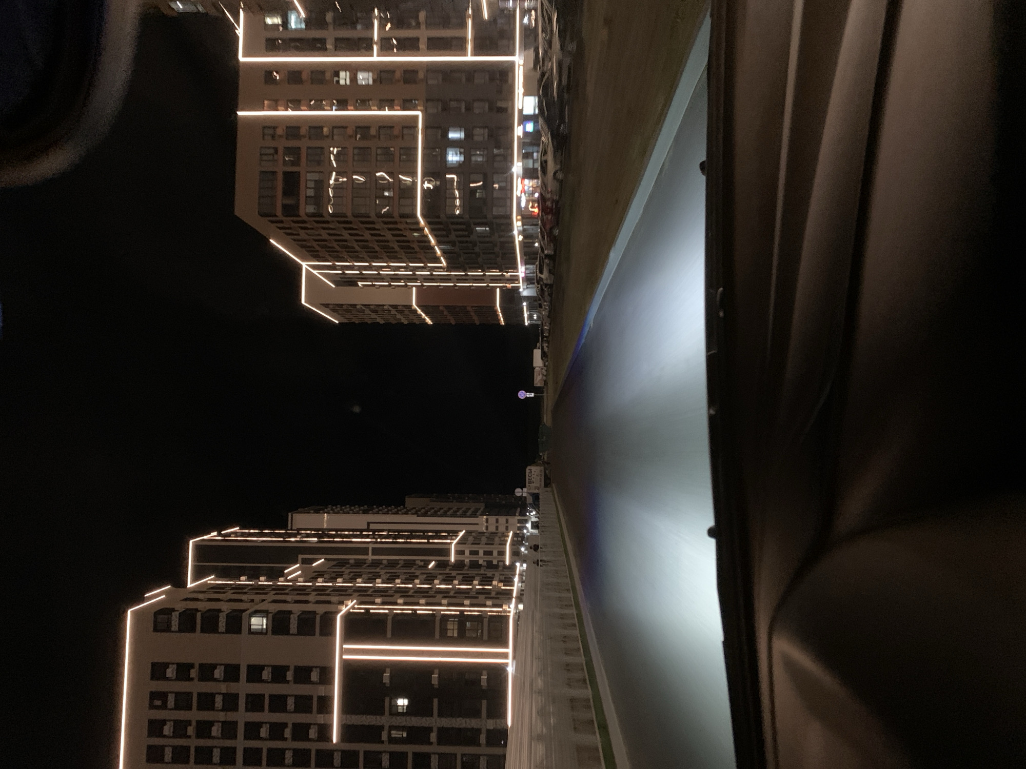 The image depicts a night-time view from the perspective of someone inside a vehicle driving on a road. The road is illuminated by the vehicle's headlights, creating a bright, blurred swath in the foreground. On either side of the road are modern, multi-story apartment buildings, their facades outlined with bright, warm-toned lights. These buildings are tall and appear to be newly constructed, with numerous windows visible. The sky is dark and black, with no stars or moon visible. The overall impression is of a contemporary urban environment at night. The buildings are identified as being located in the city of Stavropol, Russia, suggesting this is a new development or residential complex in that city. There are parked cars visible along the side of the road near the buildings. In the distance, there is a traffic sign. A few figures of people can be seen near a truck, also on the side of the road.