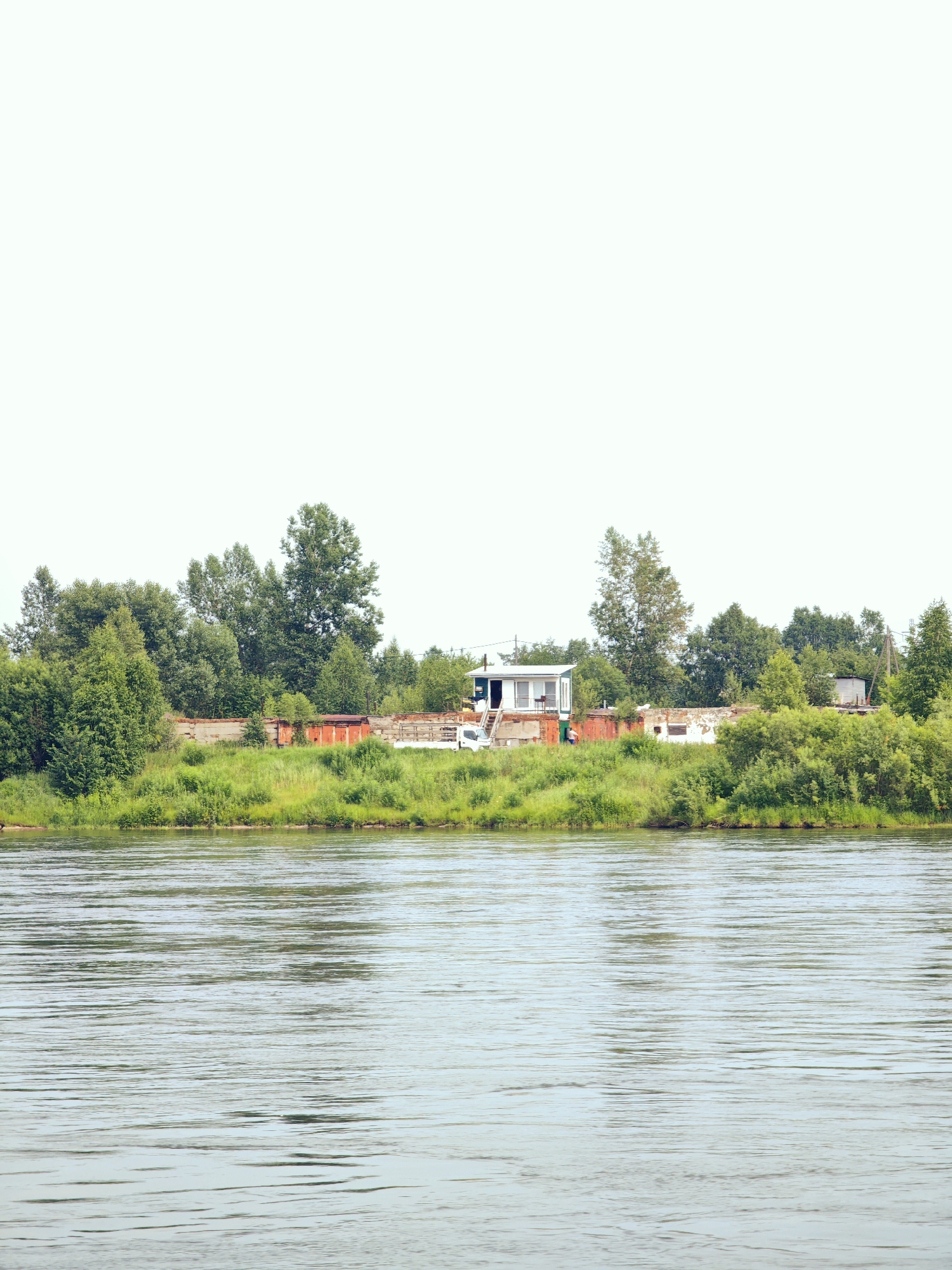 A wide, calm river fills the foreground of the image, its surface rippled by a gentle breeze. The riverbank on the opposite side is lined with lush green grass and dense trees, forming a verdant backdrop. Nestled amongst the trees are several structures, including a small white building with a blue roof and a white porch, a long, low building with red doors, and a few other smaller buildings. A white vehicle is parked near the white building, and a single person is visible near the red-doored building. The sky above is a pale, washed-out white, suggesting an overcast or hazy day. The scene appears to be a rural or semi-rural area, possibly on the outskirts of Sosnovoborsk, Russia, given its natural landscape and simple structures.