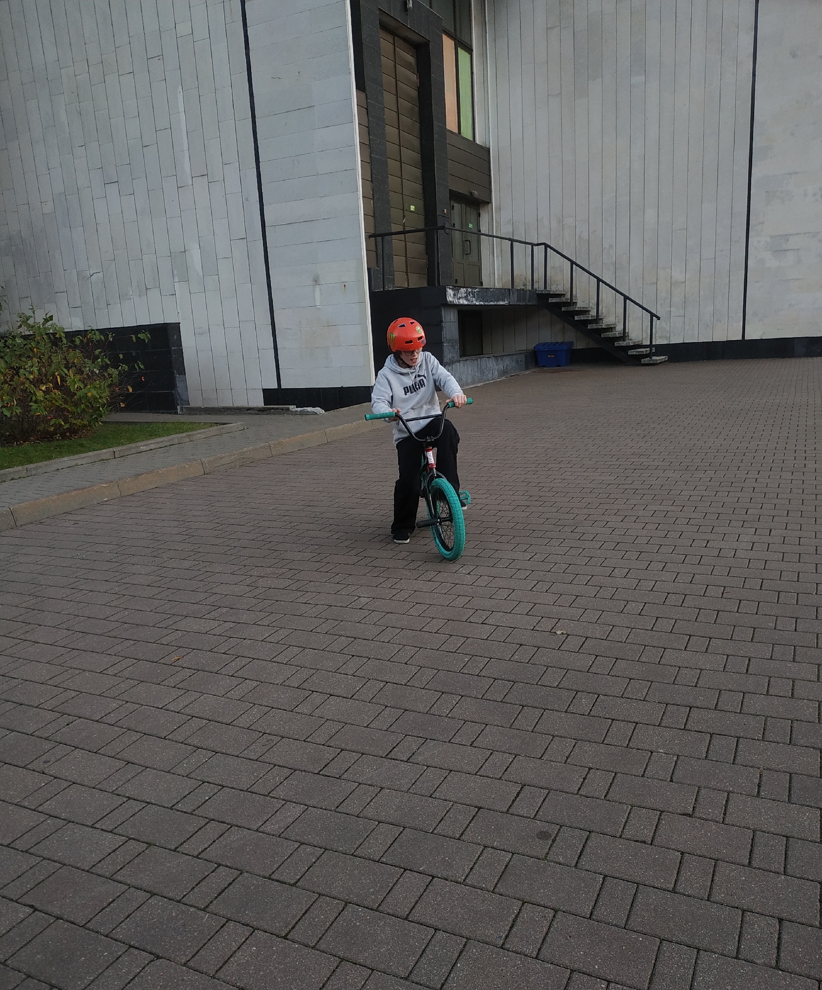 A child wearing an orange helmet and a grey Puma sweatshirt rides a turquoise-wheeled bicycle on a brick-paved area. The location is Minsk, Belarus. In the background, there is a large, multi-story building with a staircase leading to an upper entrance. A small bush is present in the lower-left corner of the frame.