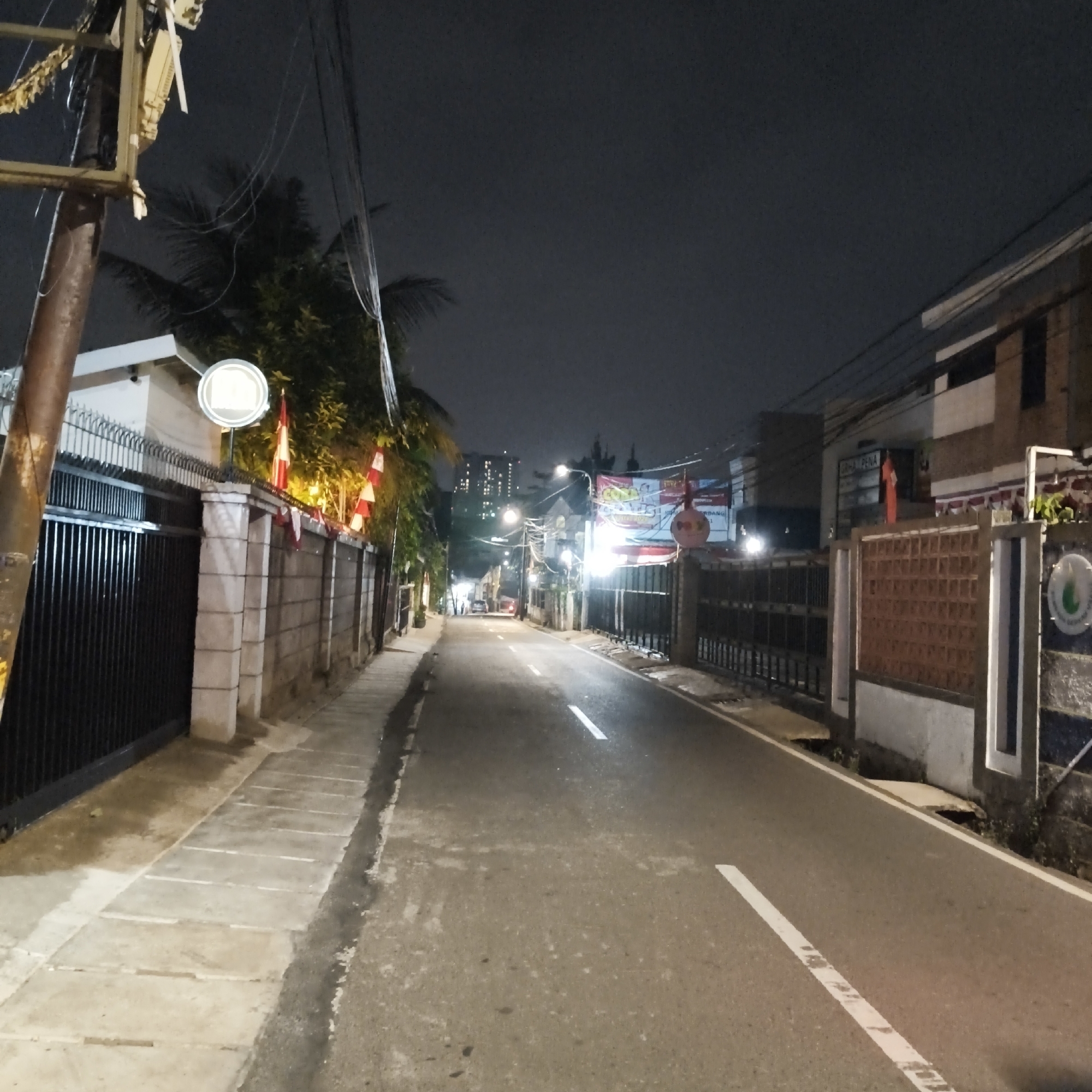 The image shows a street scene at night in South Jakarta, Indonesia. The street is paved and has white lane markings. On the left side of the street, there is a sidewalk with square paving stones and a black metal fence. Beyond the fence, there are buildings and trees. On the right side of the street, there is a wall with a patterned brick section and a white pillar. There are also buildings and fences along this side. Overhead, there are numerous electrical wires and a lamppost illuminating the street. Red and white flags are visible, attached to poles along the fence on the left side of the street and also on the right side. In the distance, a tall building with many lit windows is visible, suggesting an urban environment. Several cars are parked or driving on the street in the distance. A banner with text and graphics is hanging across the street in the mid-ground. The overall scene is dimly lit by artificial lights and the sky is dark.