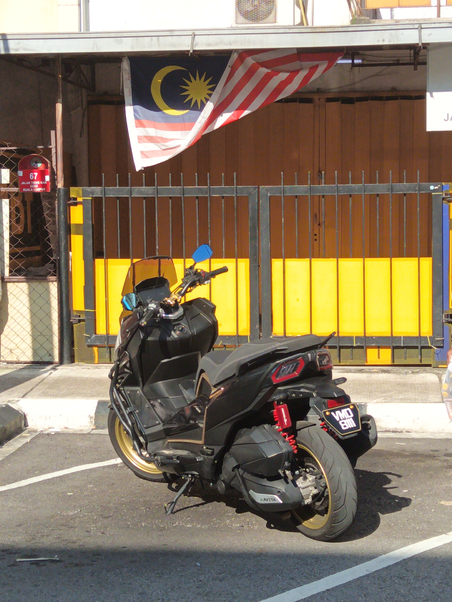 A black scooter with a gold rim and red shock absorbers is parked on asphalt in front of a building. The building has a yellow roll-up door behind a black metal gate. A Malaysian flag is hanging from the roof of the building. A red mailbox with the number 67 on it is visible to the left of the gate. The mailbox has text that reads "JALAN TRONG NABI 50350 KUALA LUMPUR." The license plate of the scooter reads "VMD 8111".