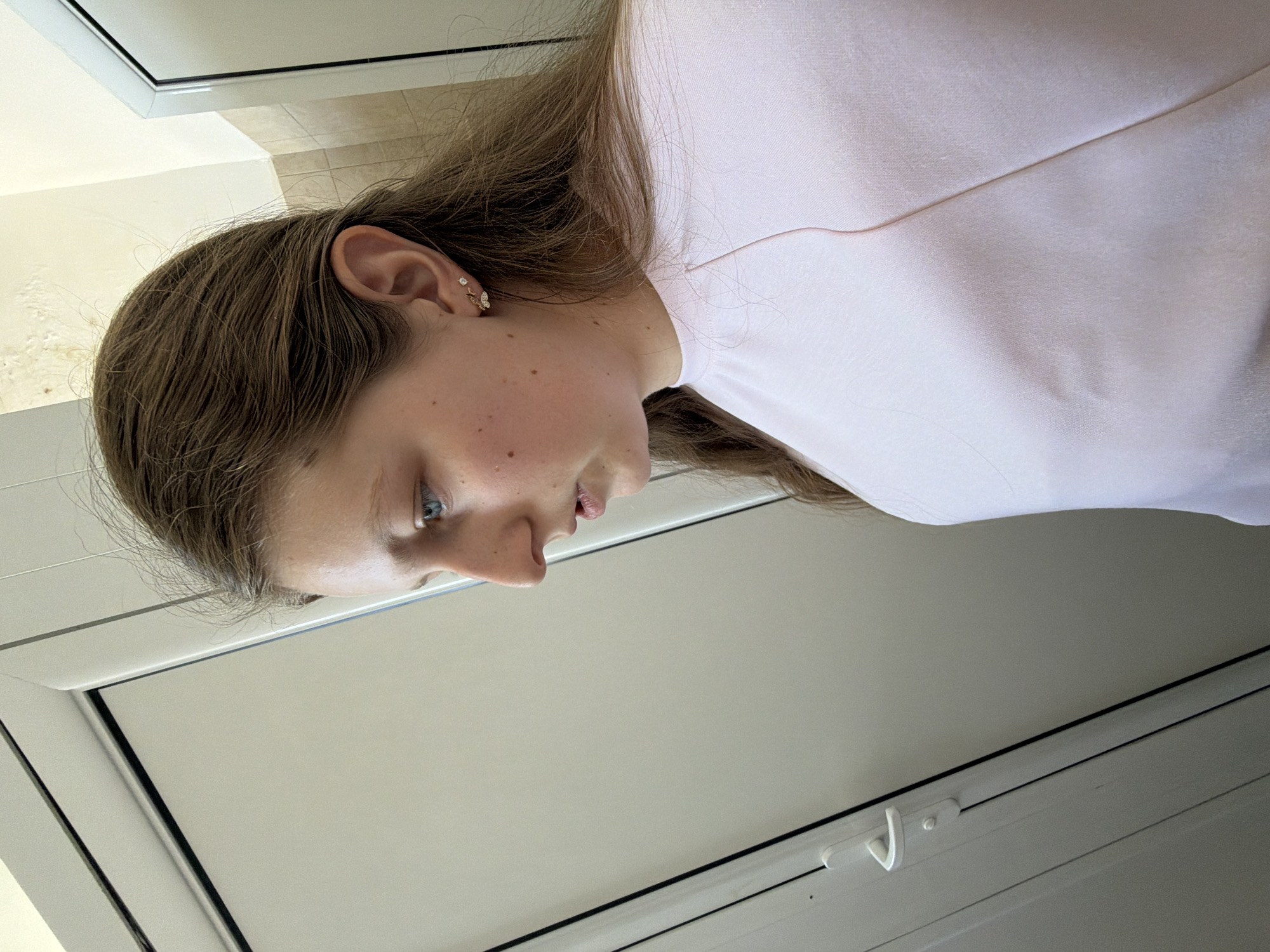 A close-up, eye-level, horizontally-oriented photograph shows a young woman indoors, seemingly gazing downward. Her complexion appears fair with some freckles. She has brown hair with an intricate earring visible in her left ear. She wears a light-colored, possibly white or pale pink, garment. The background features what looks like a white or light gray door and part of a tiled wall. The location of Burgas, Bulgaria, cannot be confirmed from this image alone.