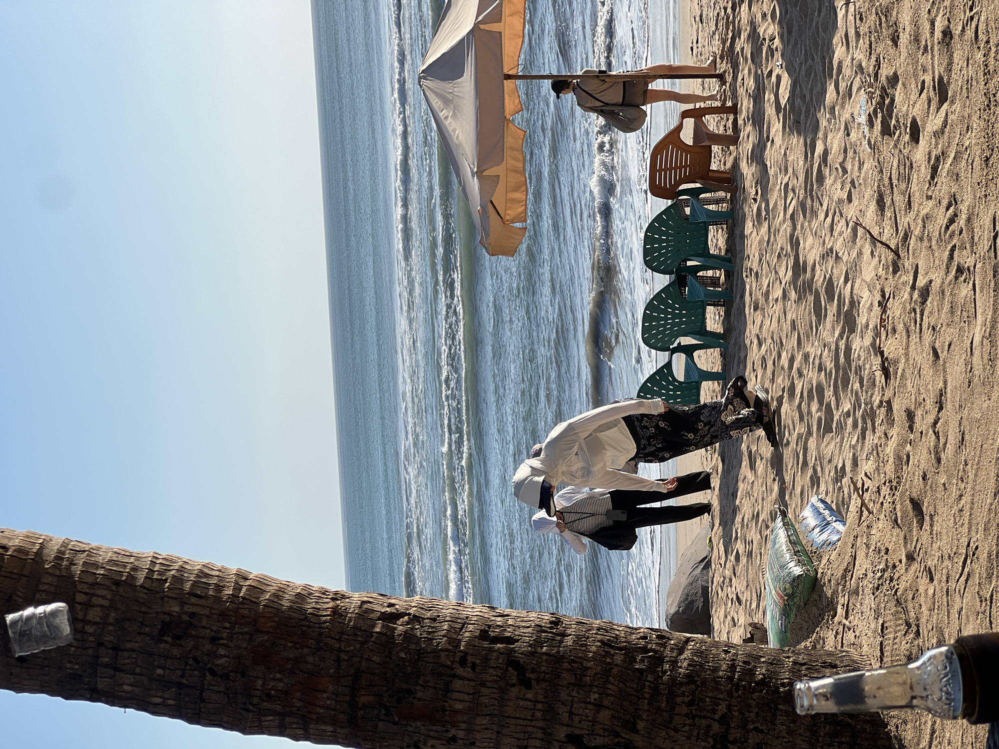 The image depicts a beach scene in Kuta, Indonesia, with the ocean in the background. A large palm tree trunk is prominently positioned on the left side of the frame, with a plastic bottle and some debris visible on the sand near its base. In the middle ground, several people are present. One individual, wearing a white long-sleeved shirt and patterned pants, appears to be walking away from the camera. To their left, two other individuals, partially obscured, are also on the beach, one wearing a white head covering and the other a striped shirt. In the foreground, there are a few plastic chairs, some colored green and one brown, arranged on the sand. An open beach umbrella stands to the right, under which a person wearing shorts and a bag slung over their shoulder is standing. The waves of the ocean are visible behind the people and chairs, with a clear blue sky above.