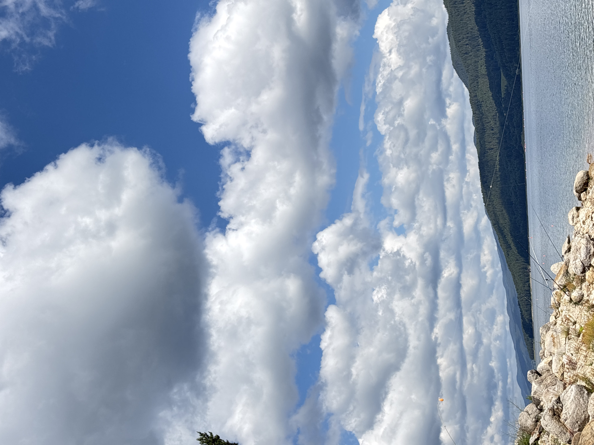 The image depicts a scenic view of a body of water, likely a lake, bordered by a rocky shoreline in the foreground and tree-covered mountains in the background. The sky is a vibrant blue, populated with a mix of fluffy white and darker, grayer clouds, suggesting a dynamic weather system. Several fishing rods are planted in the rocky bank, with their lines extending into the water, indicating recreational fishing activity. The water's surface is calm, reflecting the light and the surrounding landscape. In the distance, a faint outline of structures can be seen along the far shore, possibly indicating a small settlement or recreational facilities. A single orange float is visible on one of the fishing lines. The overall atmosphere is serene and natural, with a hint of human presence through the fishing equipment.