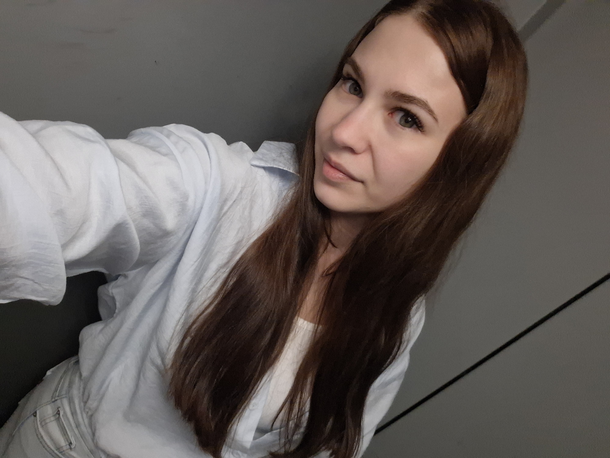 A woman with long brown hair is posing for a selfie. She is wearing a light blue button-down shirt over a white top. The background is a neutral grey wall. The location is possibly in Chaikovsky, Russia.