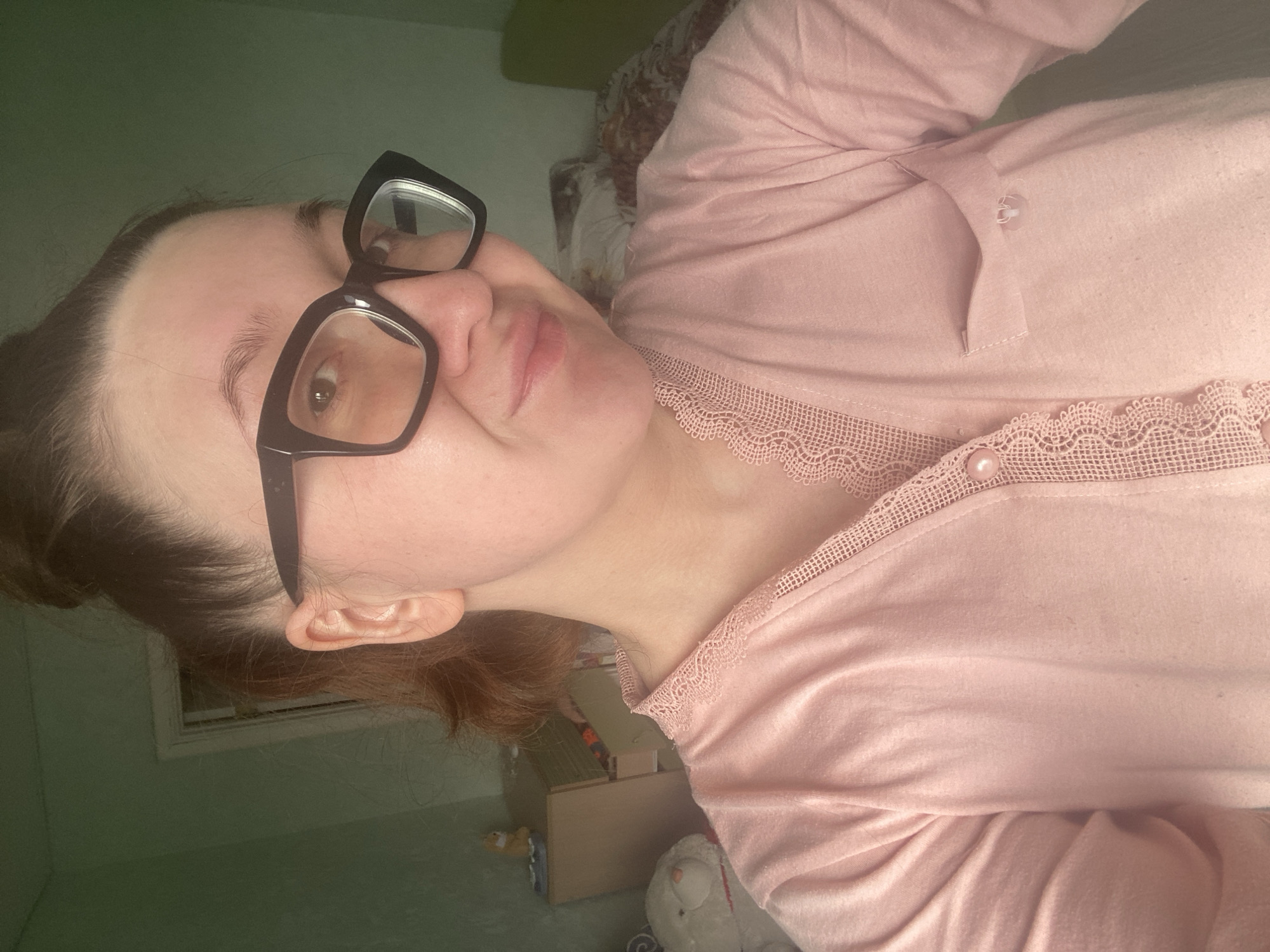 An individual, appearing to be a young adult female with dark hair pulled into a bun, is captured in a close-up indoor selfie within a residential space in Chaikovsky, Russia. She wears black-framed glasses and a long-sleeved pink top featuring lace trim along the V-neckline/placket and a pearl button. A small, open breast pocket is visible on the right side of her garment. The individual is looking towards the camera with a slight smile. The background reveals light green walls. To the left, a portion of a white door frame is visible, along with a light-colored wooden cabinet or dresser upon which a small orange figurine rests. Below the cabinet, a white stuffed animal with red accents is partially visible. To the right, a bed is present with a patterned blanket. The lighting is soft and diffused.