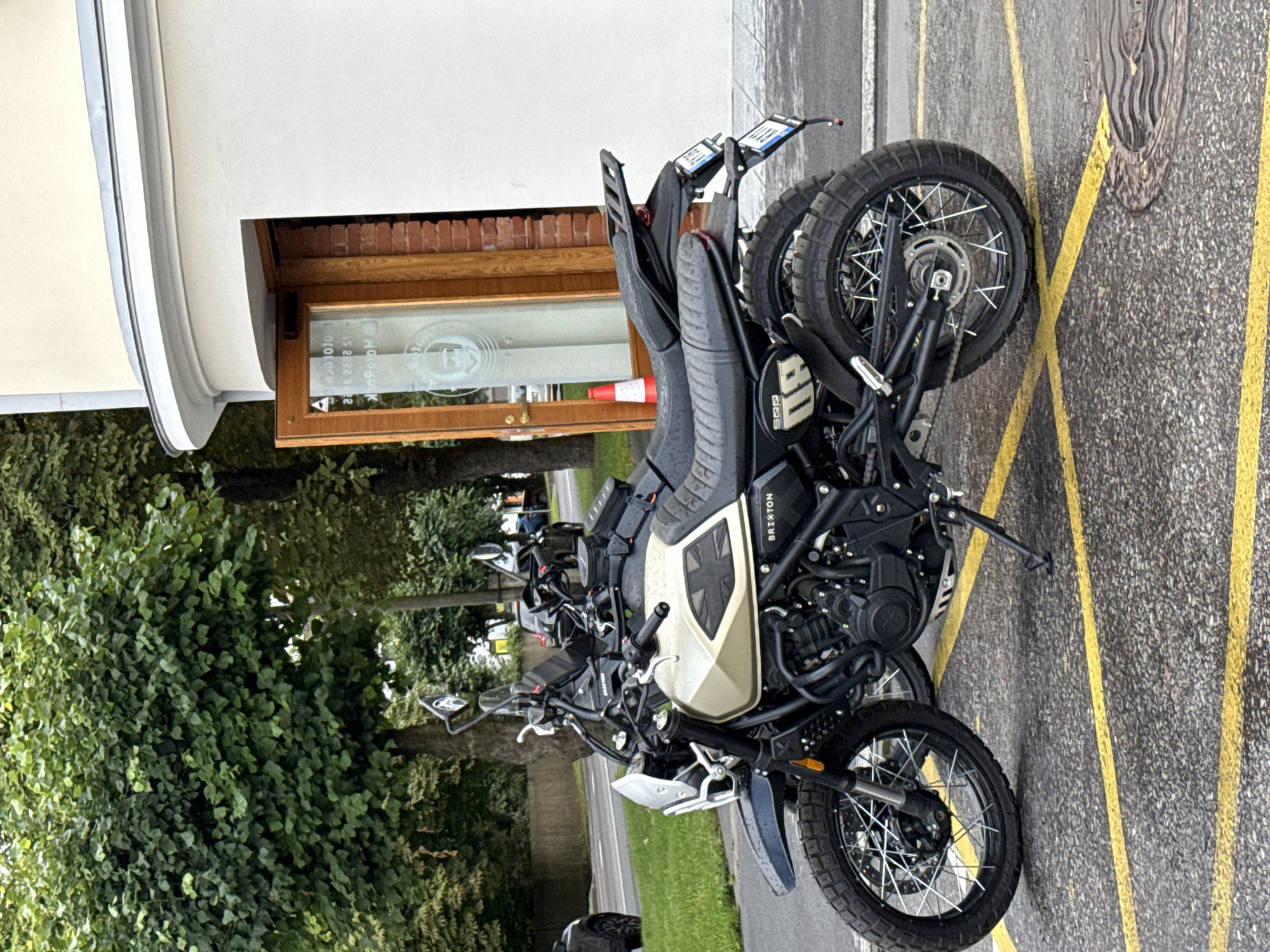 A black and tan Brixton motorcycle is parked on an asphalt surface marked with yellow parking lines, in front of a light-colored building with a wooden-framed glass door. The motorcycle is the primary subject, positioned in the foreground and angled towards the viewer's left. It has a tall windshield, spoked wheels with knobby tires, and a dark seat. The building behind it features a white facade and a curved architectural element at the top. A bright orange traffic cone is visible just inside the open doorway of the building, and a man's face is visible in a circular logo on the glass of the door. The environment appears to be outdoors in a city setting, with trees visible in the background. The motorcycle has license plates with partially visible lettering. The scene is well-lit, suggesting daytime.