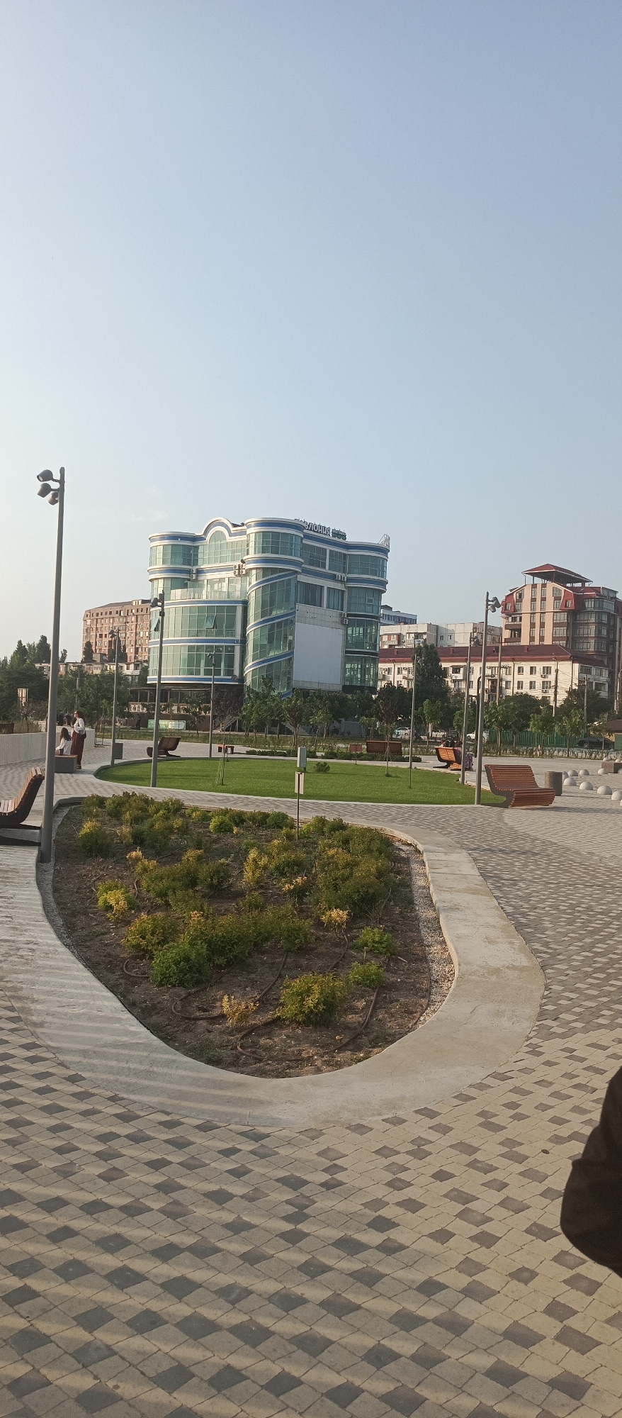 The image captures an outdoor scene in Makhachkala, Russia, on a clear day. In the foreground, a landscaped area with low green bushes and dark soil is bordered by a curved concrete pathway. The pathway is paved with interlocking diamond-shaped stones in alternating shades of grey and beige. To the left, a modern, multi-story building with a distinctive curved facade, large glass windows, and blue accents dominates the middle ground. The building has text on its upper portion that appears to read "ПРЕДЛОГИ" and "Stories". Behind this building, other urban structures, including residential buildings, are visible, some with red tile roofs.
To the left of the main building, a tall, dark grey lamp post with three lights stands. A few people are visible in the middle ground, near the lamp post and the pathway, appearing to be standing or sitting. Further back, a grassy area with trees and other benches are situated. To the right of the central building, another building with a red roof and architectural details is visible, along with more lamp posts and spherical decorative elements along the ground. The sky is a pale, clear blue. The overall impression is one of an urban public space, likely a park or promenade, with modern architecture.