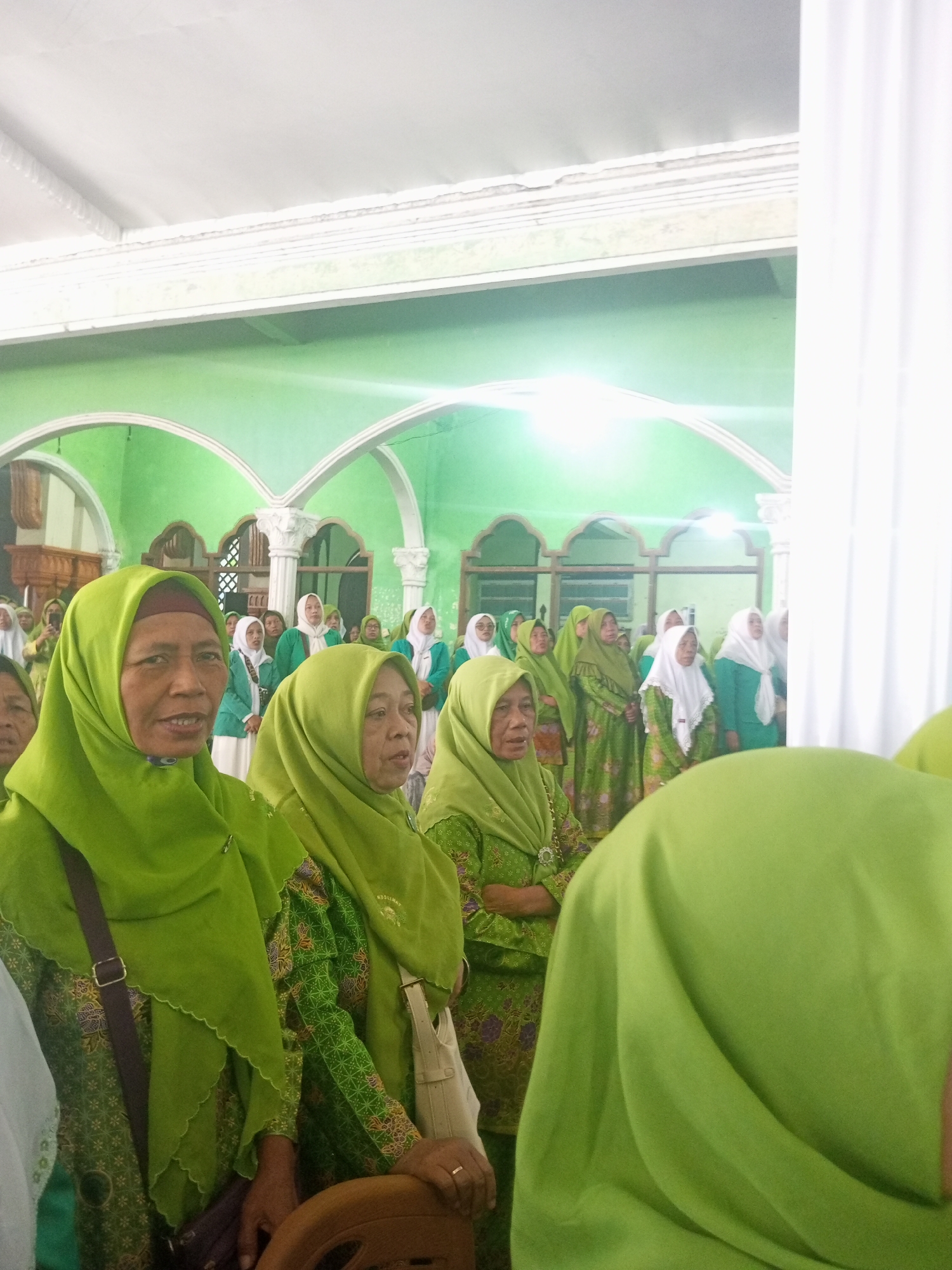The image depicts a group of Indonesian women, all wearing green hijabs and matching green patterned clothing, gathered indoors. The setting appears to be a place of worship or a community event, possibly a mosque, with arched doorways and a green-painted wall in the background. The women are standing and facing in the same direction, suggesting they are listening to a speaker or observing an event. One woman in the foreground is looking towards the camera, while others are looking off to the side. The lighting is bright, indicating an indoor space with artificial illumination. The overall atmosphere seems to be one of solemnity and community. The specific location in Klaseman, Indonesia, is not discernible from the visual information alone.