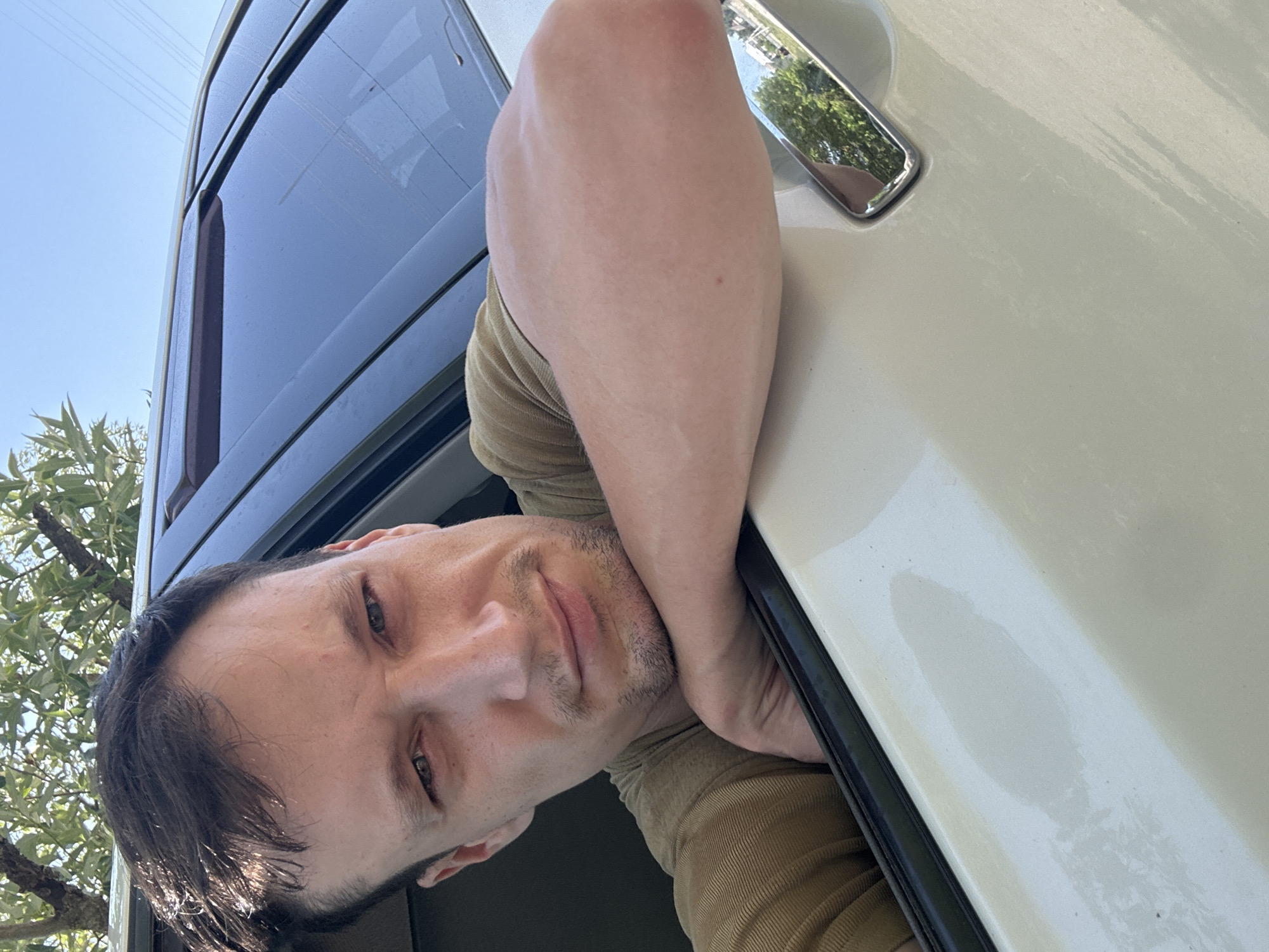 A close-up, eye-level, selfie-style shot shows a man leaning on the open window of a white car. The man has short, dark brown hair, a light complexion, and is wearing a tan t-shirt. He is looking directly at the camera with a neutral expression, and his left arm is bent and resting on the car door frame. The car's windows are tinted, and the exterior of the car is white. Trees and a blue sky are visible in the background, suggesting an outdoor setting. The image was taken in Makiv, Ukraine.