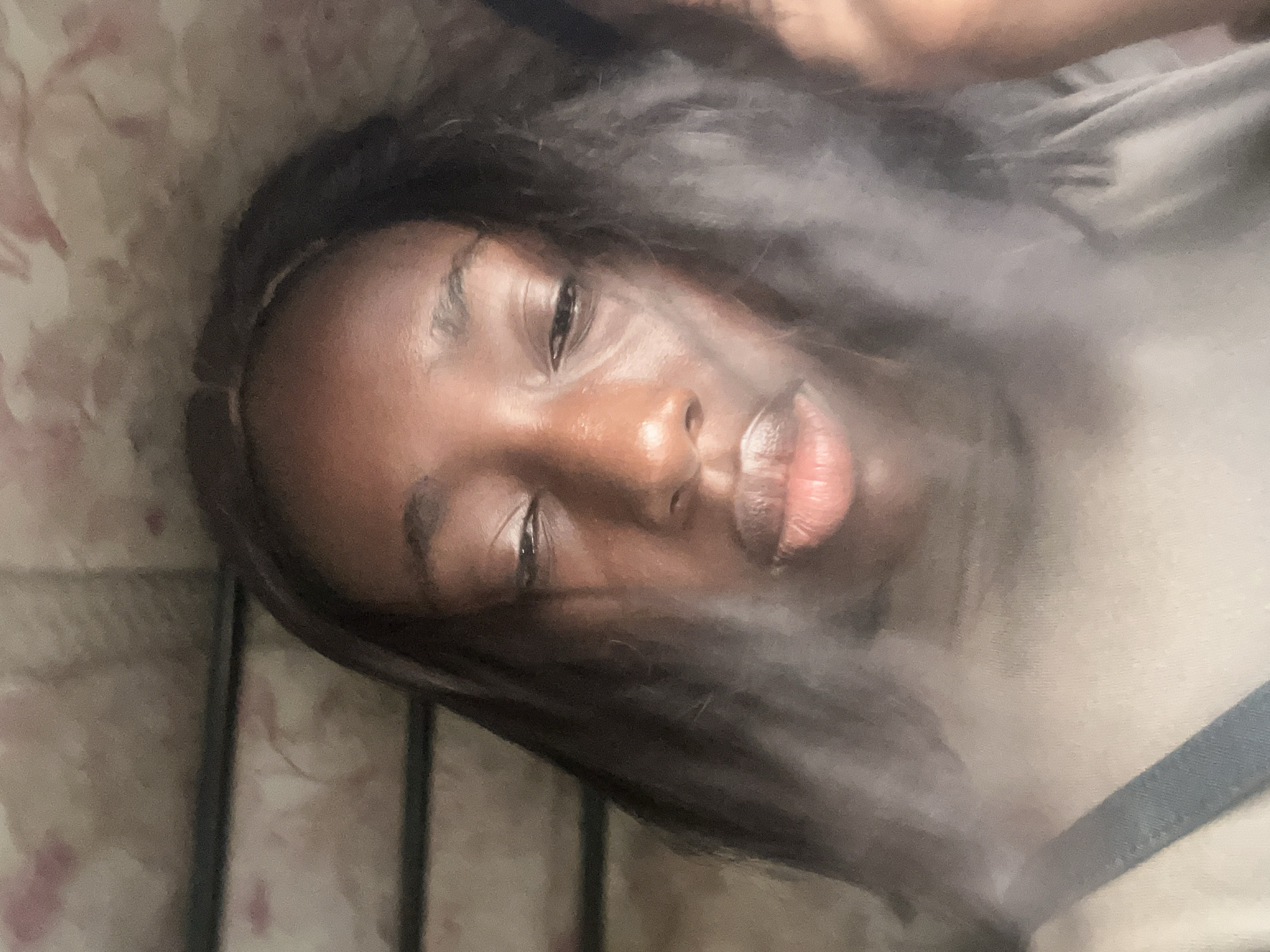A medium close-up shot depicts a Black woman lying down. Her head rests on a pillow with a light floral pattern. The woman is partially obscured by a blurry, opaque substance, possibly smoke or fog, that is concentrated in the center of the frame. She has dark hair that frames her face. Her eyes are partially closed, and her lips are slightly parted. The lighting is soft, highlighting the contours of her face. The environment is ambiguous, showing portions of bedding and a garment on the side. There is no immediate information to confirm the location is in Karum, Nigeria.