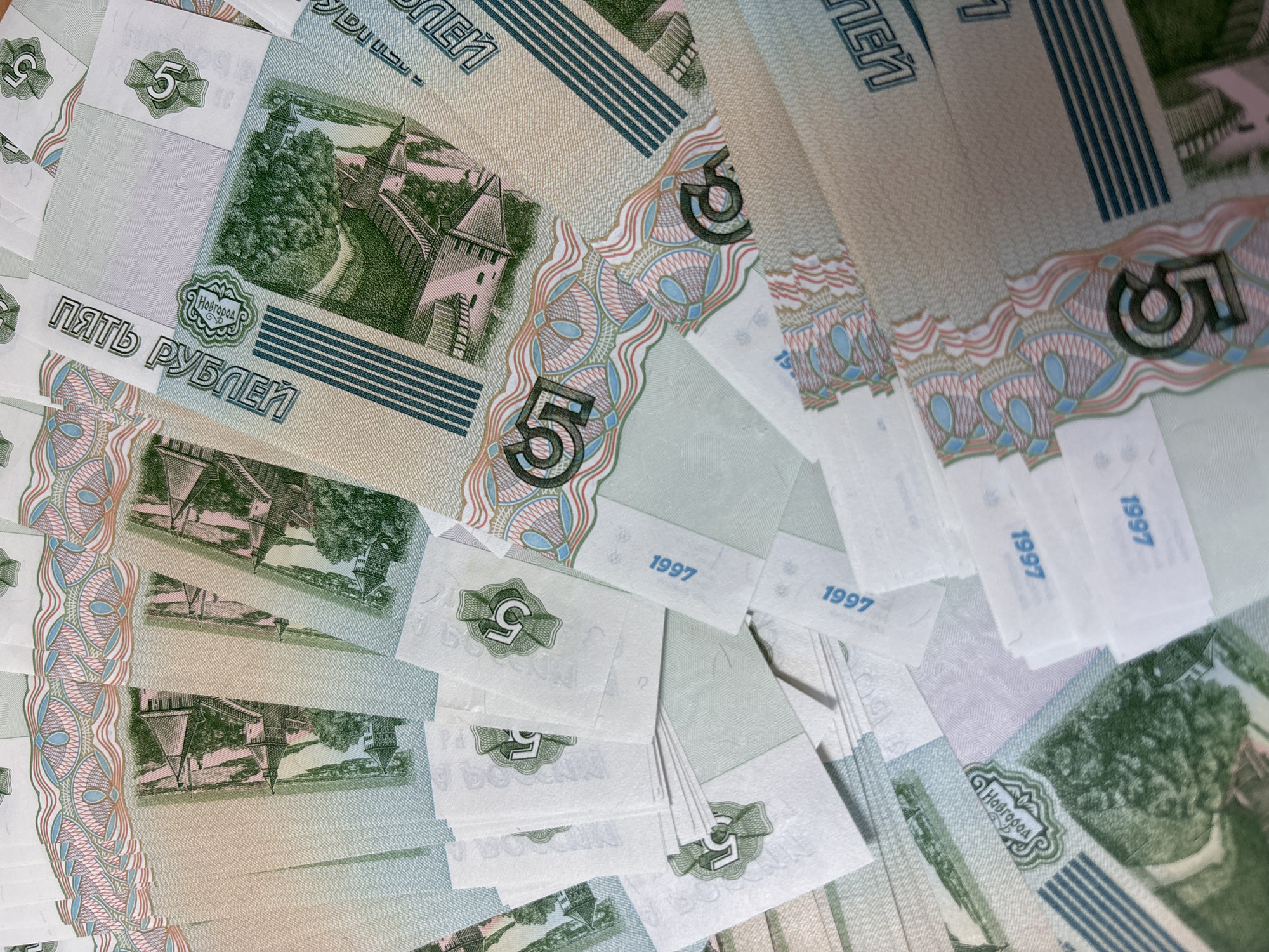 The image shows a close-up view of numerous 5 Russian Ruble banknotes. The banknotes feature the number "5" prominently and a depiction of Novgorod, a city in Russia. The year "1997" is also printed on the bills. The arrangement of the banknotes suggests they are stacked and slightly fanned out. The image is lit well, highlighting the details and patterns on the banknotes. There are no people visible in the image. The city of Perm, Russia, is not depicted on these notes.