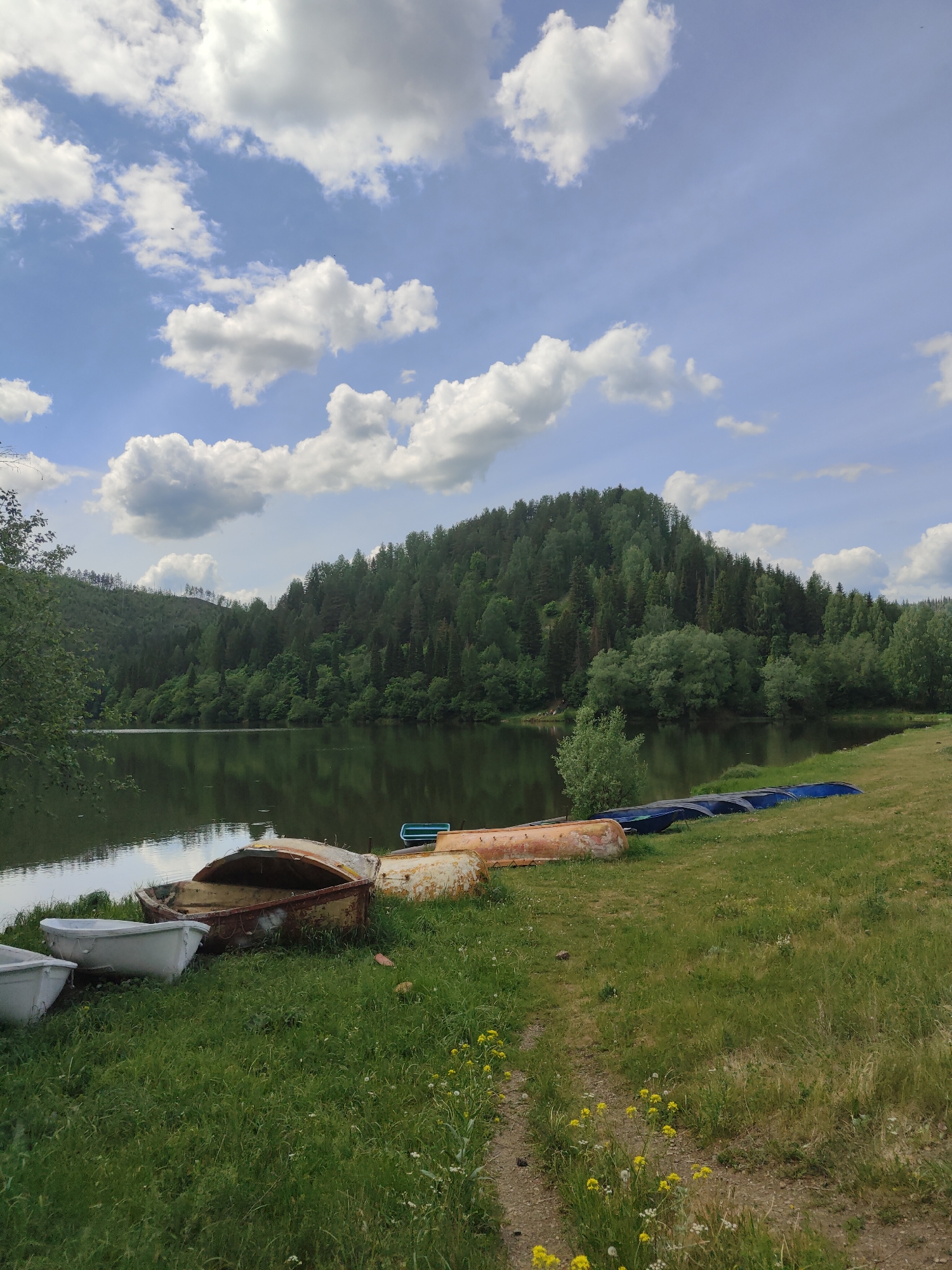 The image depicts a tranquil lakeside scene near the city of Sim, Russia. In the foreground, a grassy bank slopes down to the water's edge. Several boats, overturned and resting on the grass, are visible in varying states of repair. These include white, rusted orange, and blue boats. In the background is a still body of water reflecting the sky and surrounding vegetation. Across the lake, a forested hill rises, covered in dense greenery, and the sky is partly cloudy, with patches of blue visible.