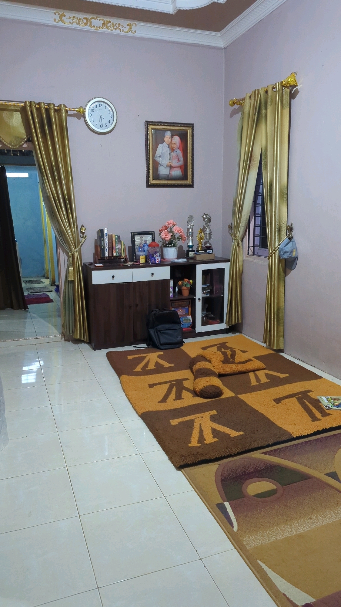 The image depicts an indoor scene, likely a living room or a study, within a residence in the city of Baroqah, Indonesia. The room is furnished with a dark wood cabinet featuring two drawers and glass-fronted compartments. On top of the cabinet, several items are displayed, including books, a framed photograph of a couple in wedding attire, a vase of pink flowers, and trophies. To the left of the cabinet, a doorway leads to another room with blue walls and two rugs visible on the floor. Golden curtains are drawn over two windows on the right and left walls. A circular clock with Roman numerals hangs on the left wall. On the floor, two patterned rugs are laid out. The first, a brown and orange checkered rug, has a cylindrical bolster pillow placed on it. A black backpack rests on the floor in front of the cabinet. The flooring is tiled in a light cream color. The lighting appears to be artificial.