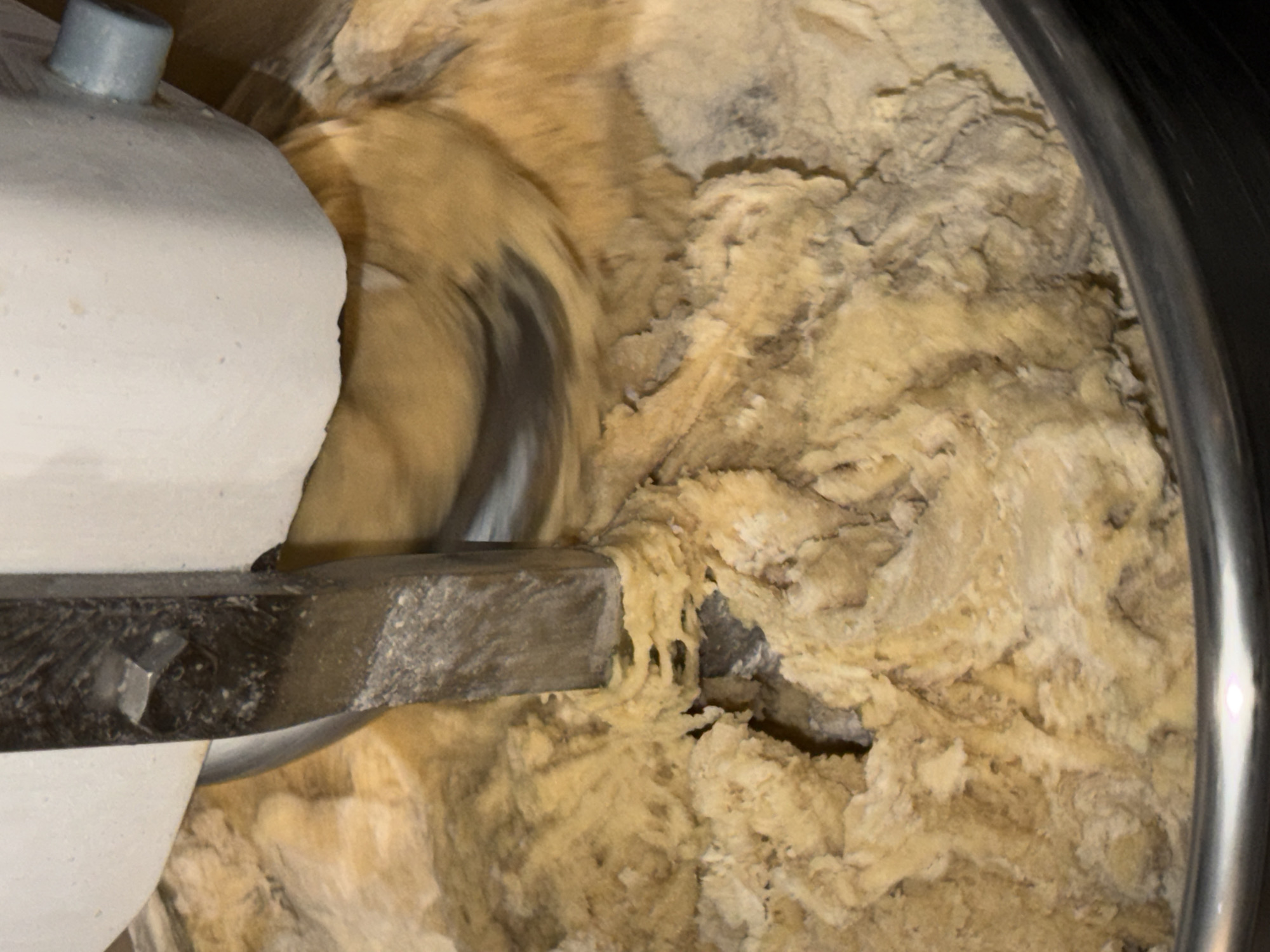 A tan, thick batter is being mixed in a large metal bowl by a metal blade attached to a white stand mixer. The mixer is on and the blade is blurred by motion. The batter is thick and clumpy, suggesting a possible dough or heavy cookie batter. The edge of the bowl is visible on the right side of the frame. The setting or location is not discernible from the provided image.