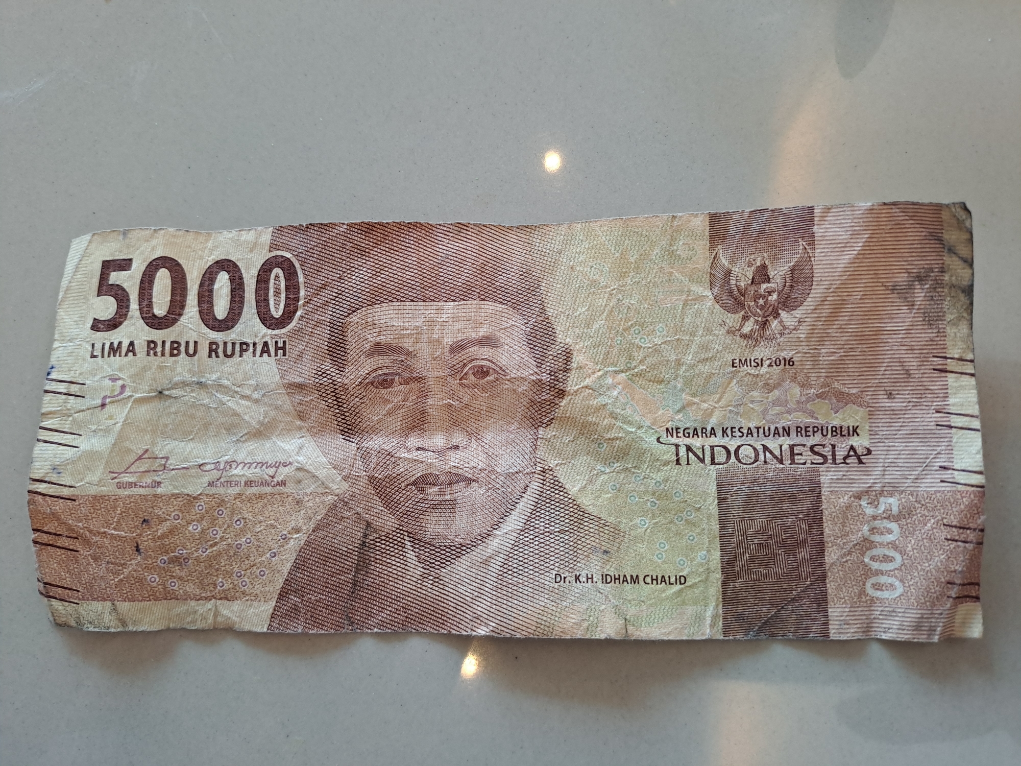 The image shows a crumpled 5000 Indonesian Rupiah banknote lying on a light-colored, textured surface. The banknote features a portrait of a man, the denomination "5000", and text in Indonesian. The banknote has visible creases and wear. The background surface appears to be a countertop or table. The image does not provide any information about the city of Bireuenmeunasahdayah, Indonesia.