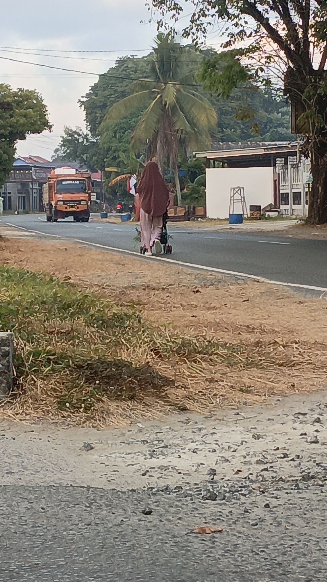 A person wearing a brown hijab and pink pants pushes a stroller along the side of a paved road in Gusunge, Indonesia. The person is walking away from the camera, and the stroller appears to be empty. The road is lined with dry grass and vegetation on the left side and trees and buildings on the right. An orange truck is parked on the left side of the road in the background, and a white building with a blue barrel and a ladder is visible on the right side. The sky is overcast.