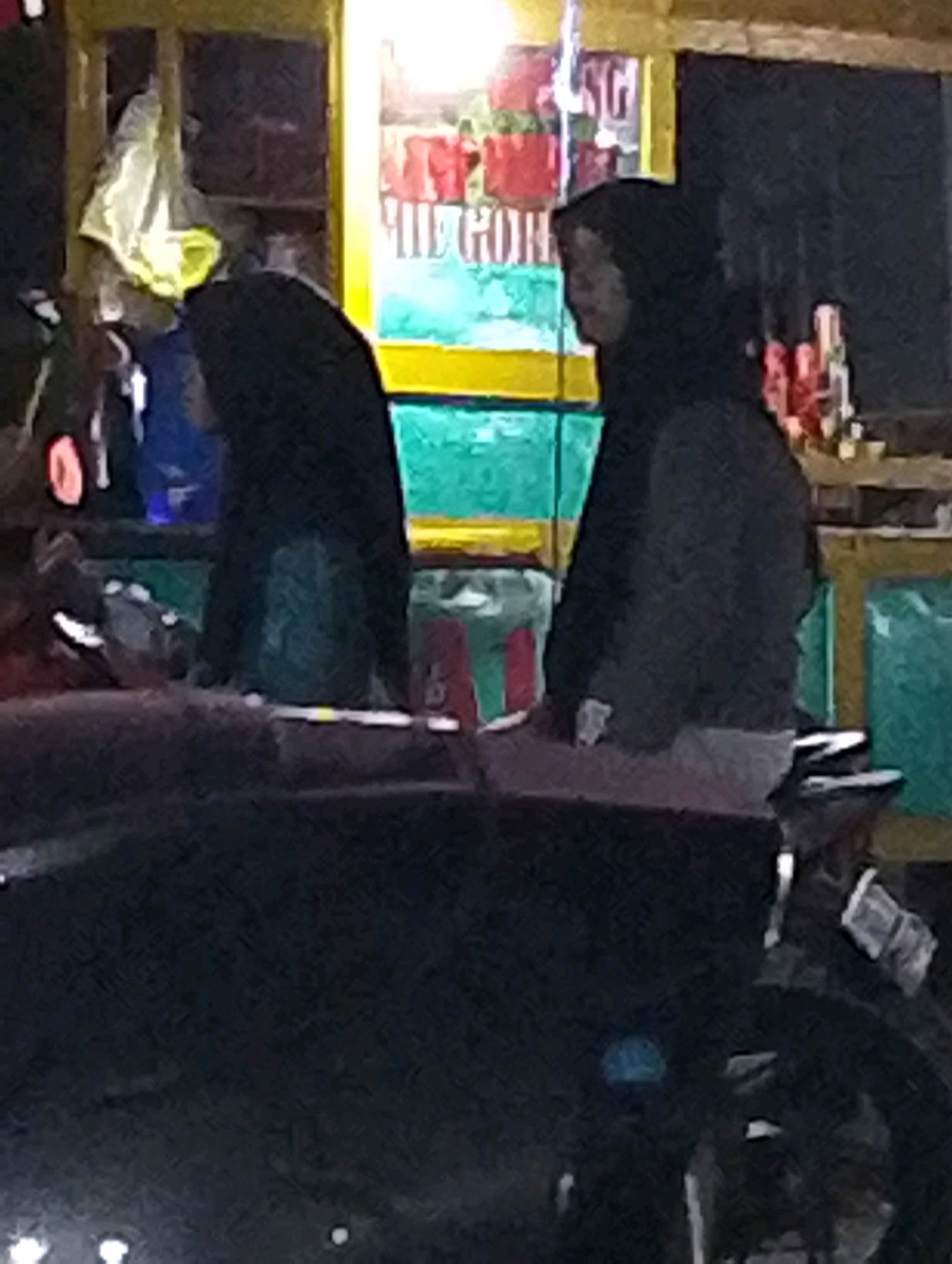 Two individuals, appearing to be women, are standing outdoors at night. The woman on the left is wearing a dark hijab and a dark garment. The woman on the right is also wearing a dark hijab and a dark jacket over a lighter-colored garment. They are positioned near a food stall or kiosk that has yellow and green paneling. In the background, there are shelves with various items, including what appear to be food products in red and green containers, and a white plastic bag hanging. The scene is dimly lit, suggesting it is nighttime. The location is identified as Bangil, Indonesia.