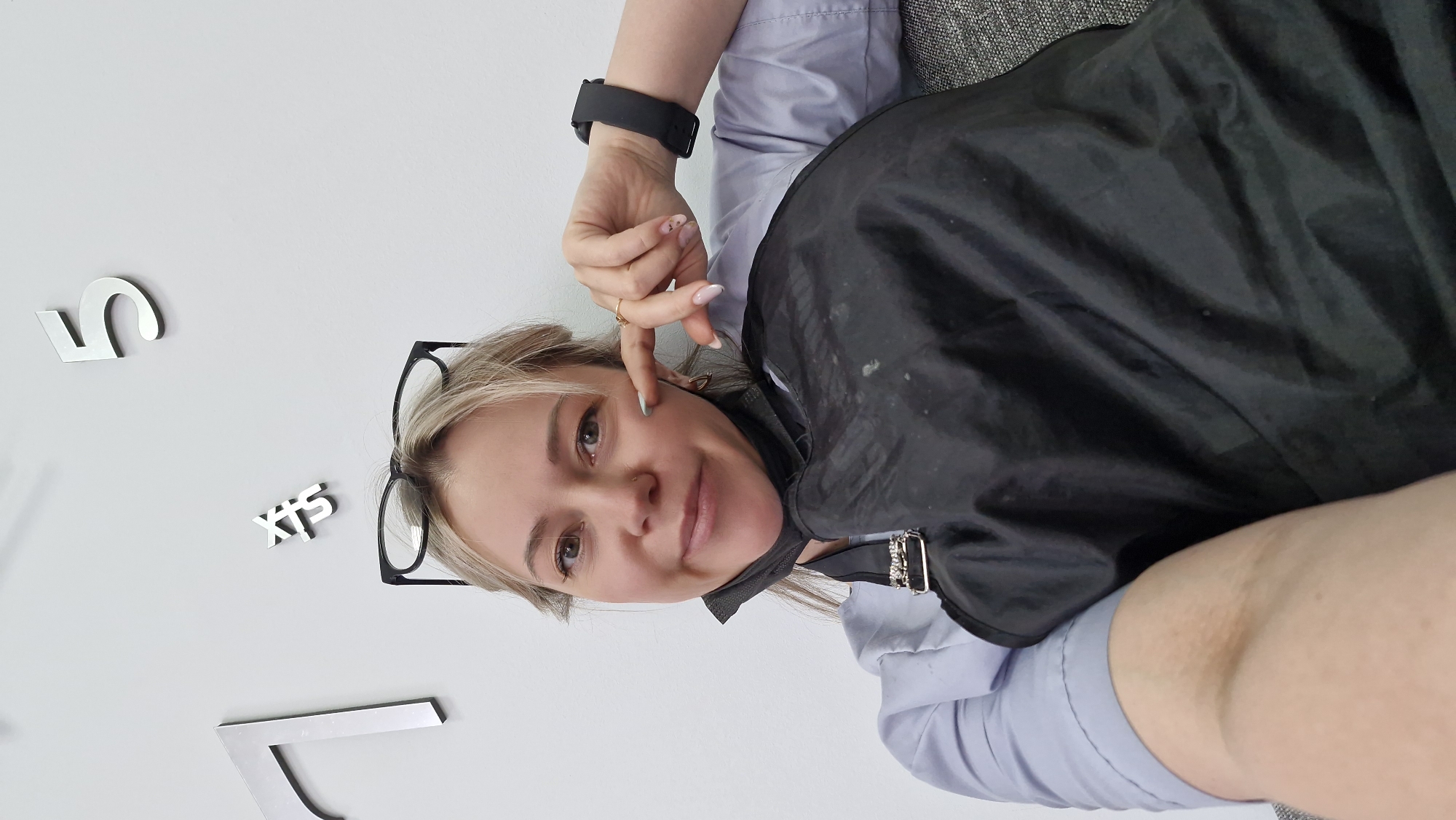 The image shows a woman posing for a selfie in an indoor environment. She has blonde hair and is wearing glasses. She is partially covered by a black salon cape, and she's wearing a light blue shirt underneath. A black smart watch is visible on her wrist, and she's wearing rings on her fingers. Her hand is near her face, touching her cheek. The background features a white wall with silver numbers and letters arranged to form a clock, possibly indicating the time. Based on the metadata, the location is Otchor, Russia.