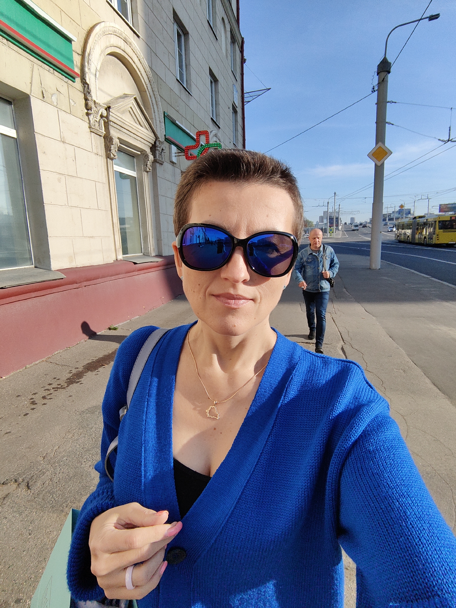 A woman with short hair and sunglasses is taking a selfie on a sunny day. She is wearing a blue cardigan over a black top and a gold necklace with a charm. A man in a denim jacket is walking on the sidewalk behind her. In the background, there is a street with cars and a yellow tram. The building on the left appears to be a commercial establishment with green signage. The scene is likely set in a city, possibly Minsk, Belarus, given the architectural style and the presence of a tram.