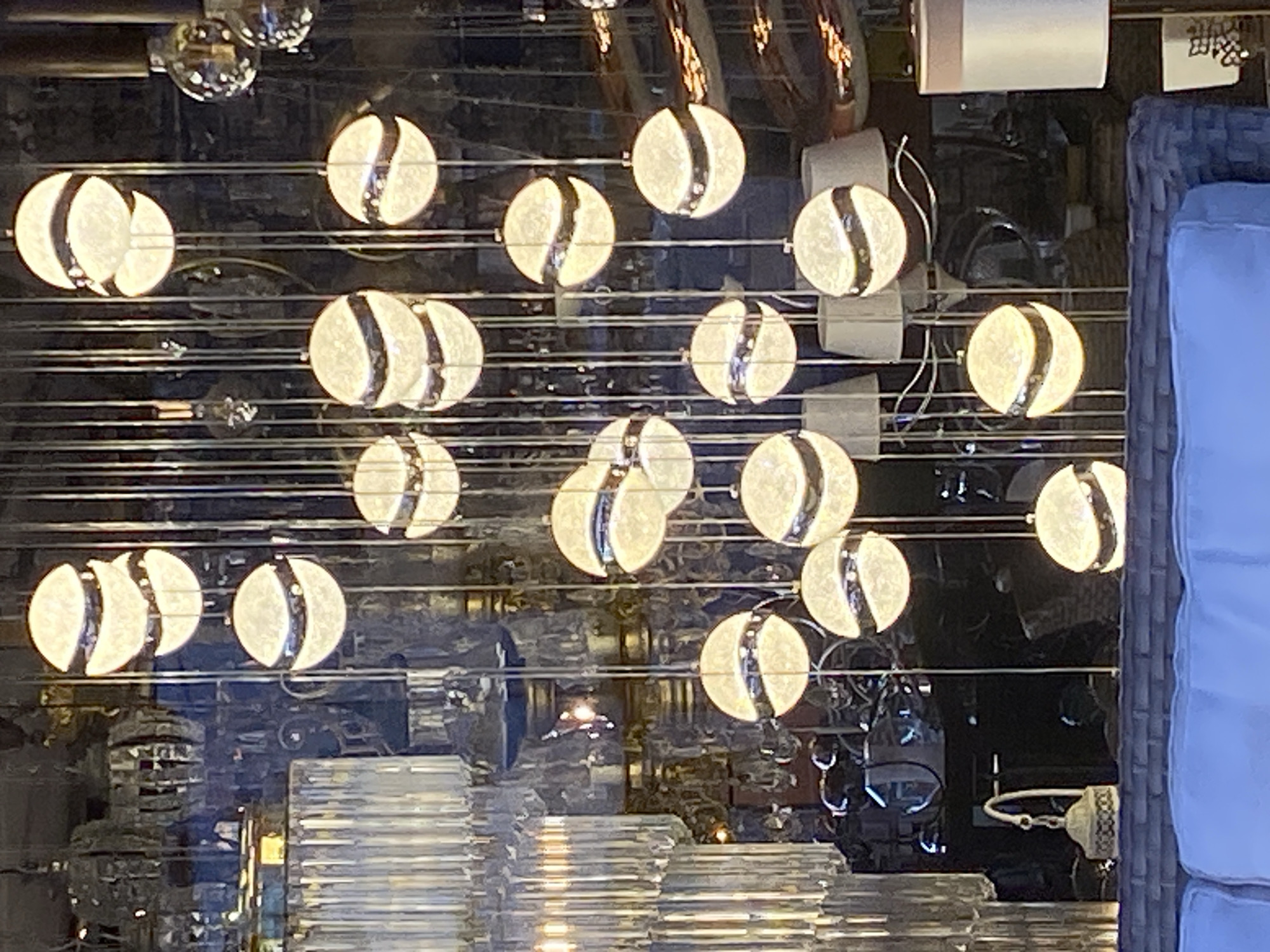 The image captures a display window likely in a commercial setting, possibly in Alanya, Turkey. Reflected in the glass are various light fixtures. There are spherical bulbs with a band of light across the center. The window also reflects stacked translucent objects, suggesting a display of glassware or decorative items. The setting seems urban due to the complexity of the reflected scene. A portion of furniture, potentially an outdoor chair with a blue cushion, is visible on the right edge. The general atmosphere is shiny and modern.