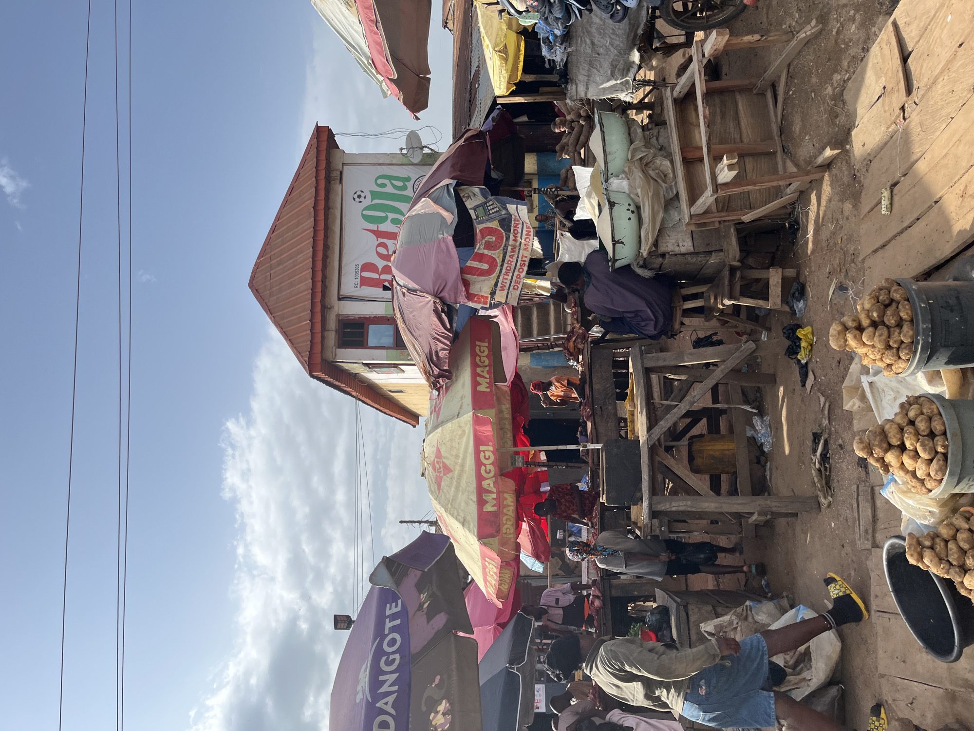 The image depicts an outdoor market scene under a clear sky with scattered clouds, likely in Rafin Par, Nigeria. Several stalls are set up, covered by colorful umbrellas, indicating a bustling marketplace. In the background, a building with a red-tiled roof features a prominent banner for "Bet9ja," a sports betting company.
The foreground and middle ground show various market activities. People are seen tending to stalls, interacting with customers, and moving around. There are piles of produce, possibly potatoes, displayed in buckets and on wooden platforms. Other items visible include sacks, wooden tables, and what appear to be raw meats at some stalls. The ground is dirt and scattered with debris, typical of an outdoor market. The overall atmosphere suggests a lively commercial environment.