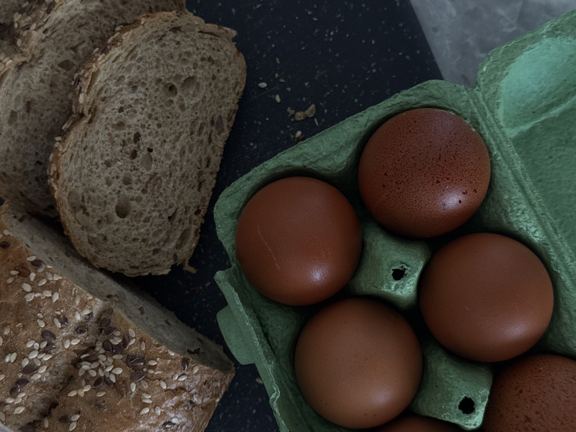 The image depicts a still life arrangement consisting of sliced bread and brown eggs in a green carton. The bread appears to be multi-grain and contains seeds on the crust. The eggs are contained within a green cardboard egg carton. The background is dark and speckled with breadcrumbs. There is no indication of this photo being taken in Saratov, Russia, or any city, for that matter. There are no people depicted in this image.