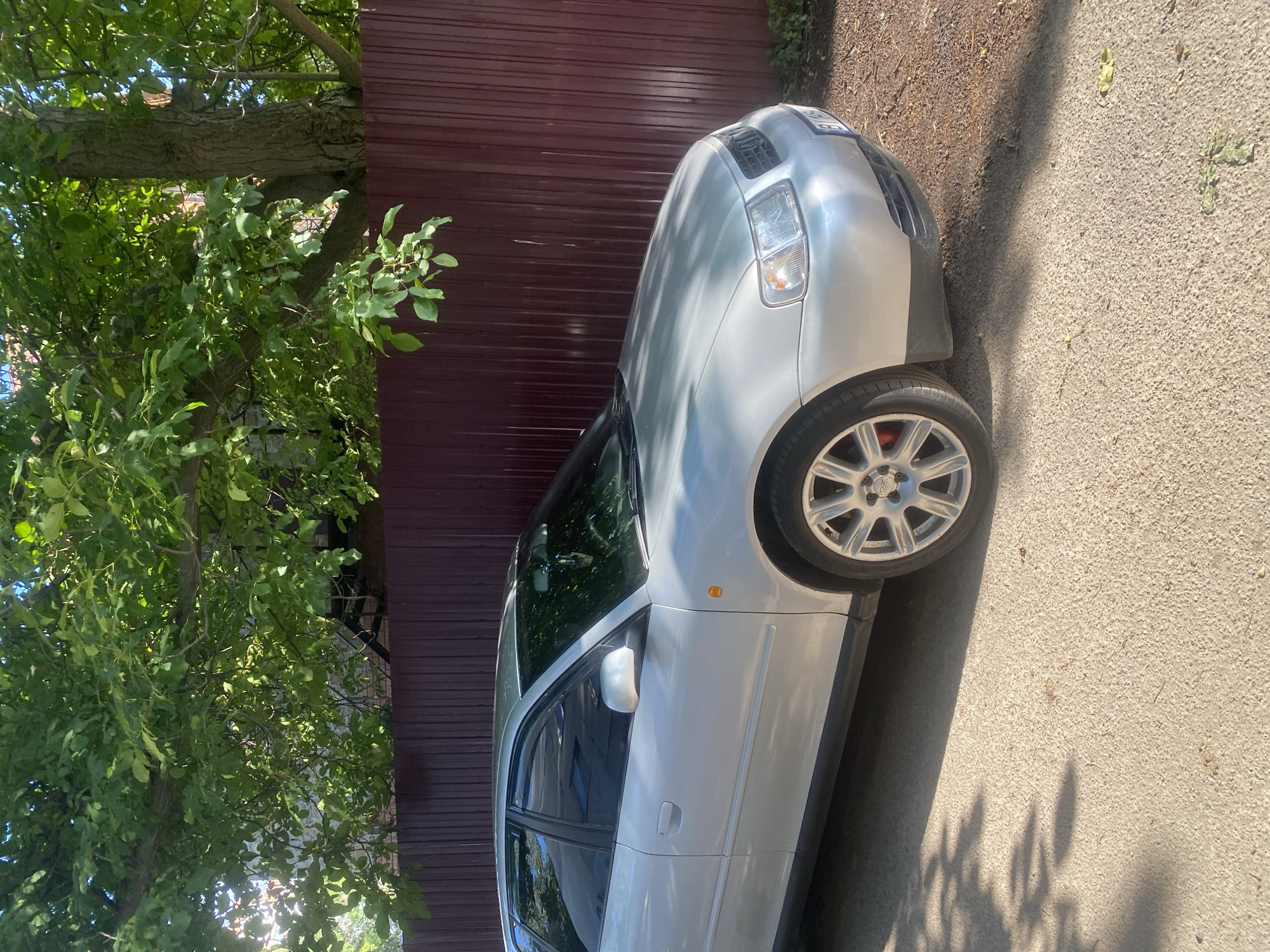 A silver Audi A3 hatchback is parked on a gravel driveway next to a dark red corrugated metal fence. Lush green tree branches overhang the scene, casting dappled shadows on the car and ground. The car is facing right, with its front quarter visible. The ambient light suggests it is daytime. The location is identified as Poltava, Ukraine, based on the context provided. There are no people or animals present in the image.