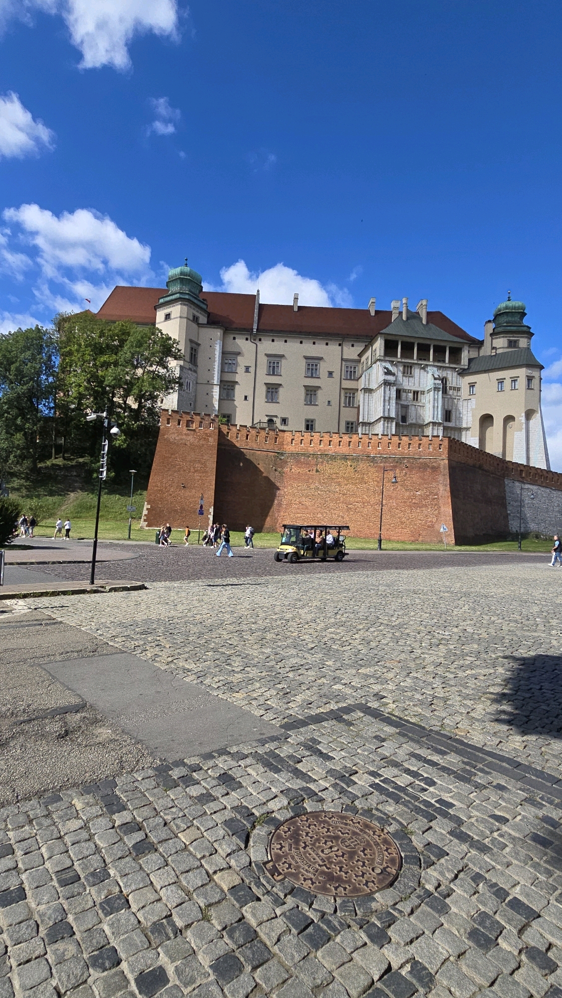 The image depicts a sunny day in Krakow, Poland, likely at Wawel Castle, given the architectural style and the prominent brick wall. The foreground is paved with cobblestones and features a manhole cover. In the middle ground, a yellow tourist vehicle with passengers is driving on a paved area. Beyond the vehicle, a tall, weathered brick wall surrounds the castle grounds. Several people are walking on the grass and paved areas in front of the wall. The castle itself, with its distinctive red-tiled roofs and green-domed towers, stands majestically on a hill in the background under a bright blue sky with scattered white clouds. Trees line the left side of the castle.