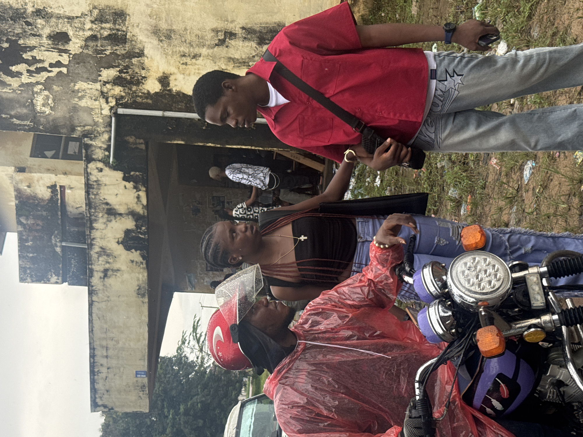 Three young adults are gathered near a motorcycle and a dilapidated building. The person on the left, wearing a red rain jacket and a red helmet, is seated on a motorcycle with purple accents. A young woman stands to the right of the motorcyclist, dressed in a black top and ripped jeans, with her hair in braids. Another young man is on the far right, wearing a red button-down shirt, a white undershirt, and light-colored jeans, with a black bag slung across his chest. In the background, other individuals are visible near the entrance of a weathered, two-story building with peeling paint. The ground around them is covered in dry grass and scattered litter. The environment appears to be outdoors, possibly in a residential or commercial area in Ihiagwa, Nigeria. The lighting suggests an overcast day.
