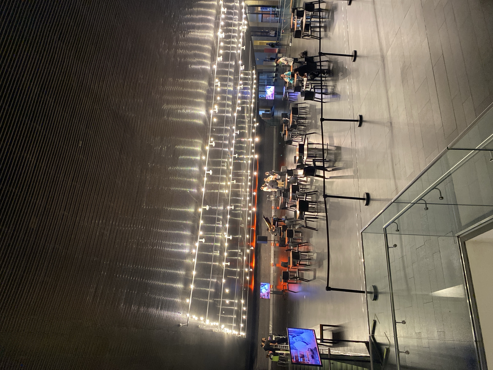 The image shows an outdoor dining area, likely in Buenos Aires, Argentina. Tables and chairs are arranged along a wall adorned with strings of lights. Several people are seated at the tables. Barriers and monitors are strategically placed to separate the dining section. The location features a wall, lighting, and possibly part of a building. The picture is taken from an elevated perspective.