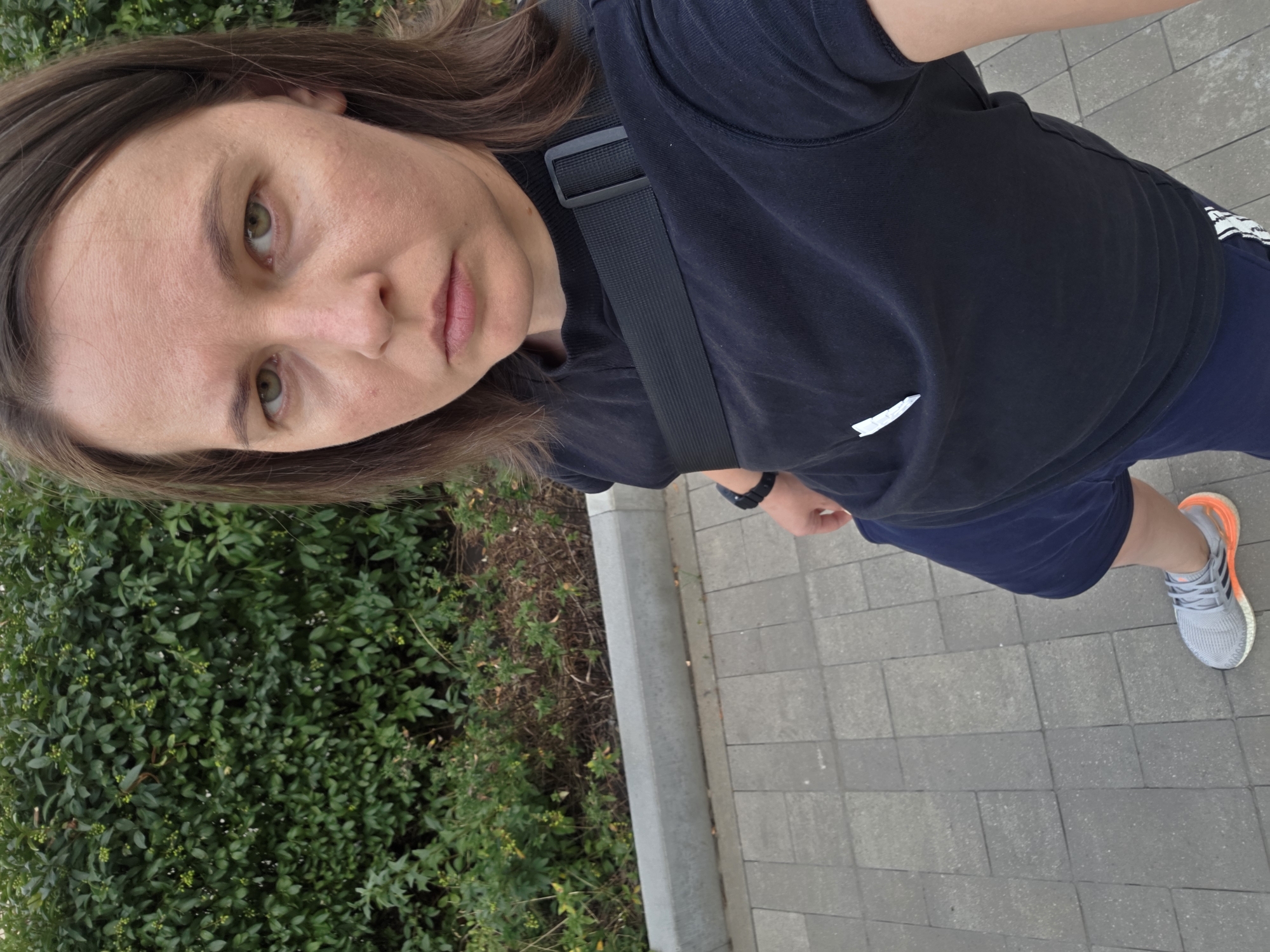 A medium shot taken outdoors shows a person with brown hair and green eyes looking directly at the camera. They are wearing a black t-shirt, dark blue shorts, and grey athletic shoes with orange accents. A black strap from a bag is visible across their chest. The person is standing on a paved walkway, with a grey concrete curb and a dense green hedge behind them. The scene is consistent with an outdoor public space in Rostov-on-Don, Russia, possibly a park or a street lined with greenery.