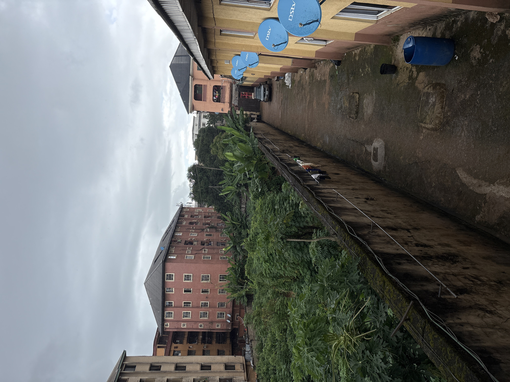 This is an outdoor, medium-angle shot of a narrow alleyway in Onitsha, Nigeria. The scene is overcast, with a gray sky overhead. The alleyway is bordered by a tall, weathered concrete wall on the left and a two-story yellow and pink building on the right. The wall on the left is covered in moss and has wires running along it. Clothes are hanging on a line attached to the wall.
The alleyway floor is paved with concrete and shows signs of wear and tear, with some cracks and patches. Several square concrete structures, possibly manhole covers, are visible on the ground. A large blue plastic barrel stands near the right wall, along with a smaller black bucket.
In the background, several multi-story buildings are visible, including a pink apartment building with multiple balconies and windows, and a taller beige building to its left. A dark-colored SUV is parked in the alleyway. Lush green vegetation, including what appear to be banana trees and other dense foliage, grows behind the wall on the left. Several blue satellite dishes with the "DSTV" logo are mounted on the right building. The overall impression is one of a densely populated urban environment.