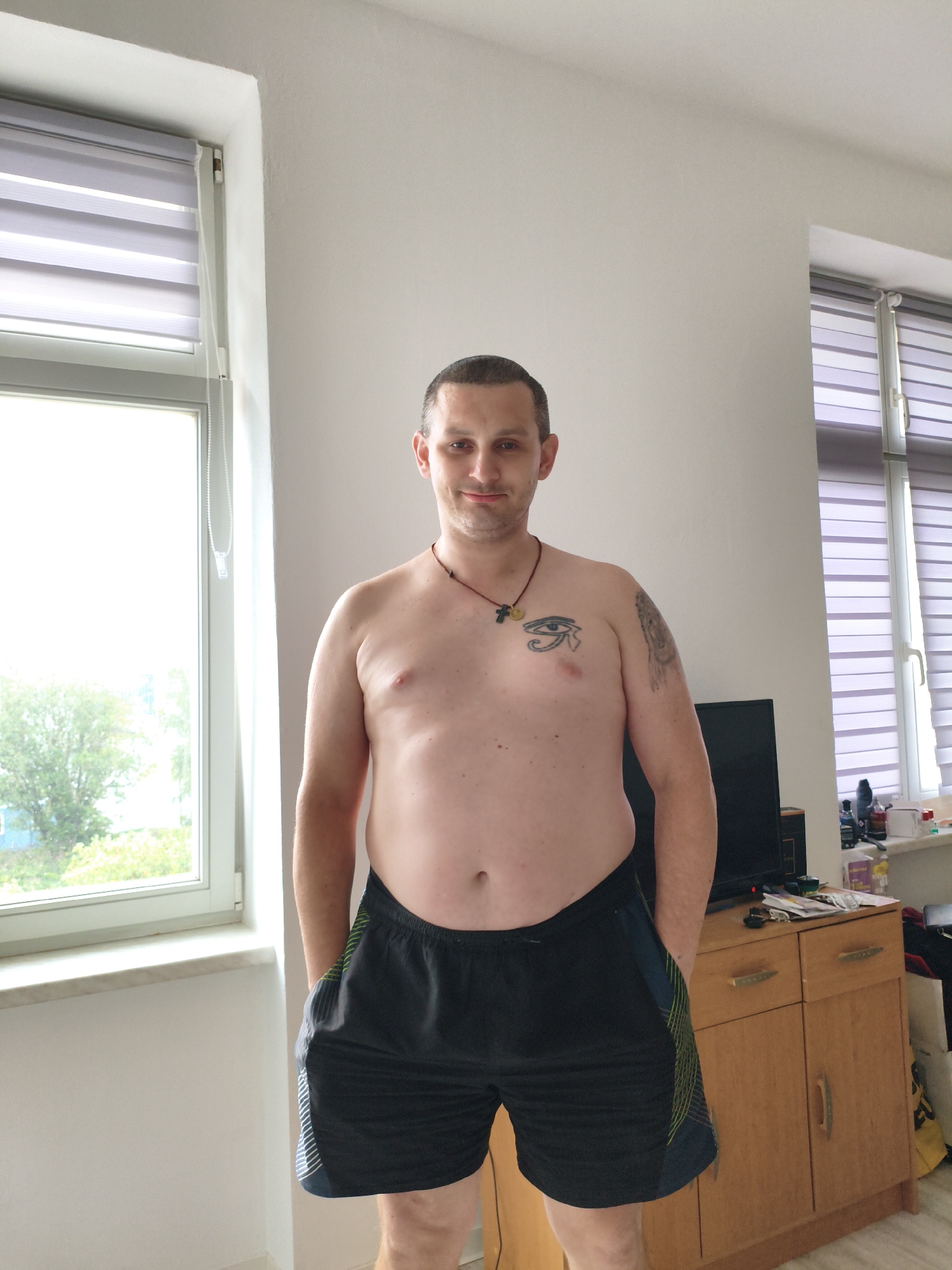 A man stands shirtless in black athletic shorts in a room with white walls and two windows covered by blinds. The man has short brown hair and is looking directly at the camera with a slight smile. He has a tattoo of an eye on his chest and a necklace with a pendant. The room appears to be a bedroom or living area, with a wooden dresser and a television visible in the background. The windows offer a view of trees and a building. This image was taken in Wolgast, Germany.