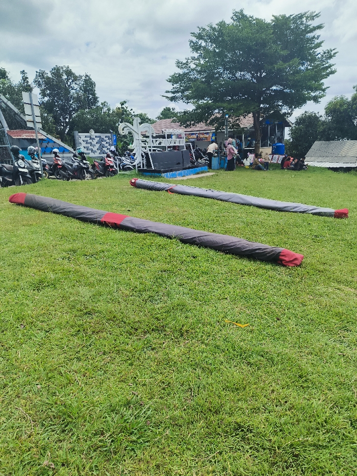 The image shows two long, black, cylindrical objects, possibly rolled-up tarpaulins, lying on a grassy field in Majalengka, Indonesia. The field is green and well-maintained. The objects are lying on the grass, with the top object slightly closer to the viewer. The background shows a few people in the distance, some standing and some sitting, near a building and a few parked motorcycles. There are several trees in the background, including a large, leafy tree in the center. The coordinates provided suggest the location is in the rural area of Majalengka, away from the main city center. The scene appears calm and peaceful, with no indication of any activity or interaction around the rolled-up tarpaulins.